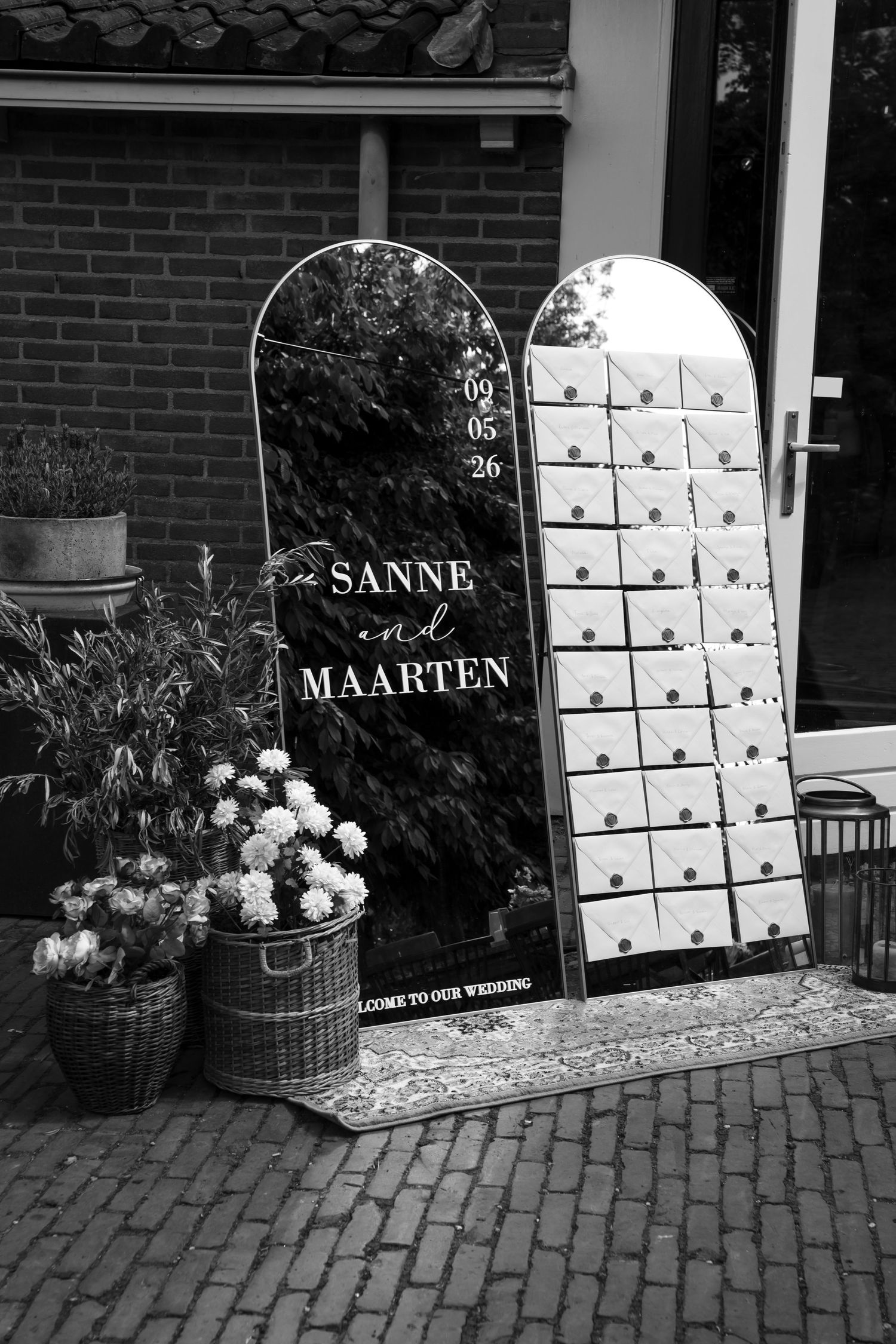 Black and white wedding welcome sign for Sanne and Maarten dated 09/05/26, displayed outdoors with floral arrangements.