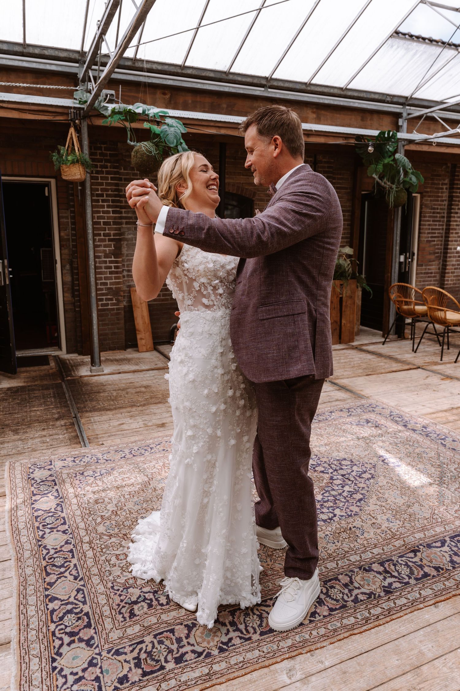 Bride and groom share first dance in elegant greenhouse venue, bride in floral white gown, groom in mauve suit.