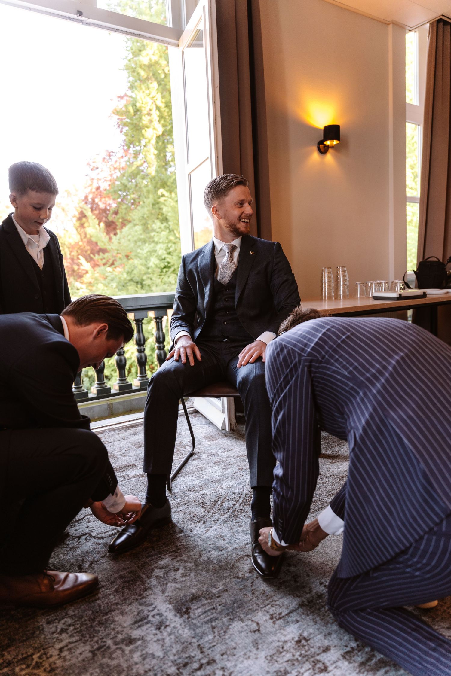 Groomsmen helping a smiling groom put on his dress shoes before a wedding ceremony in an elegant room.