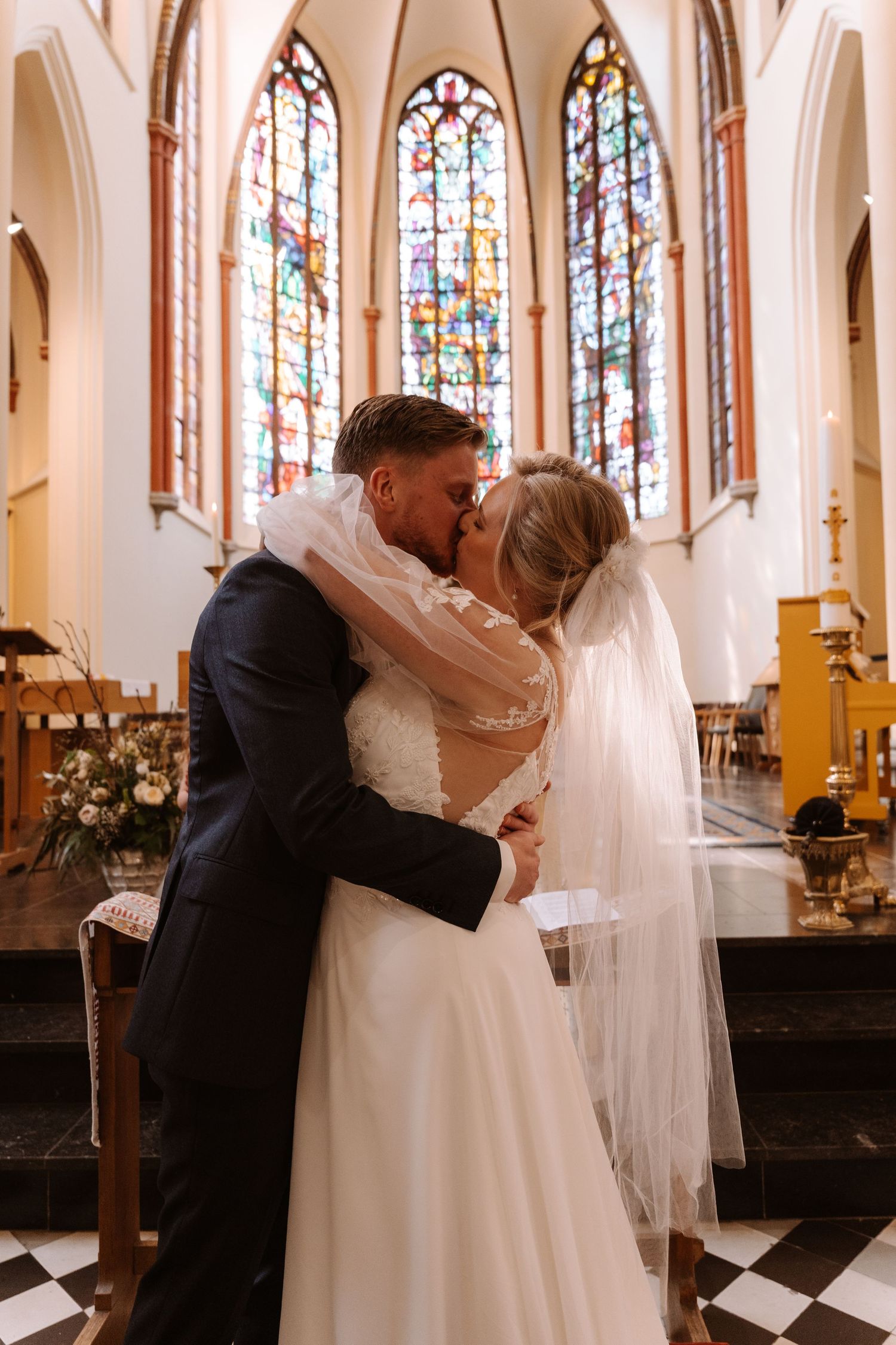 Bride and groom share first kiss at church altar with stained glass windows during wedding ceremony.