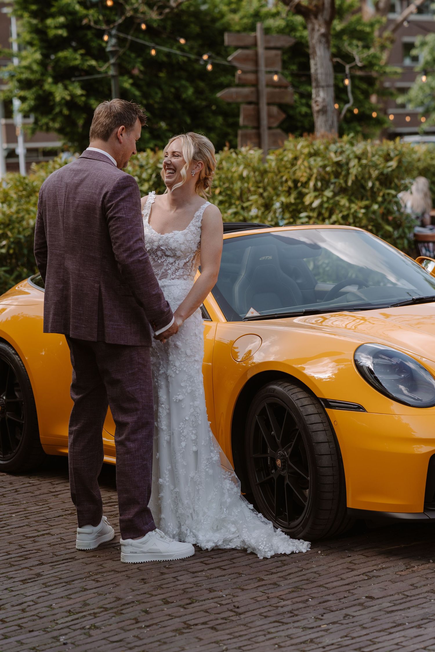 Bride and groom laughing together beside a bright yellow Porsche sports car on a cobblestone driveway.