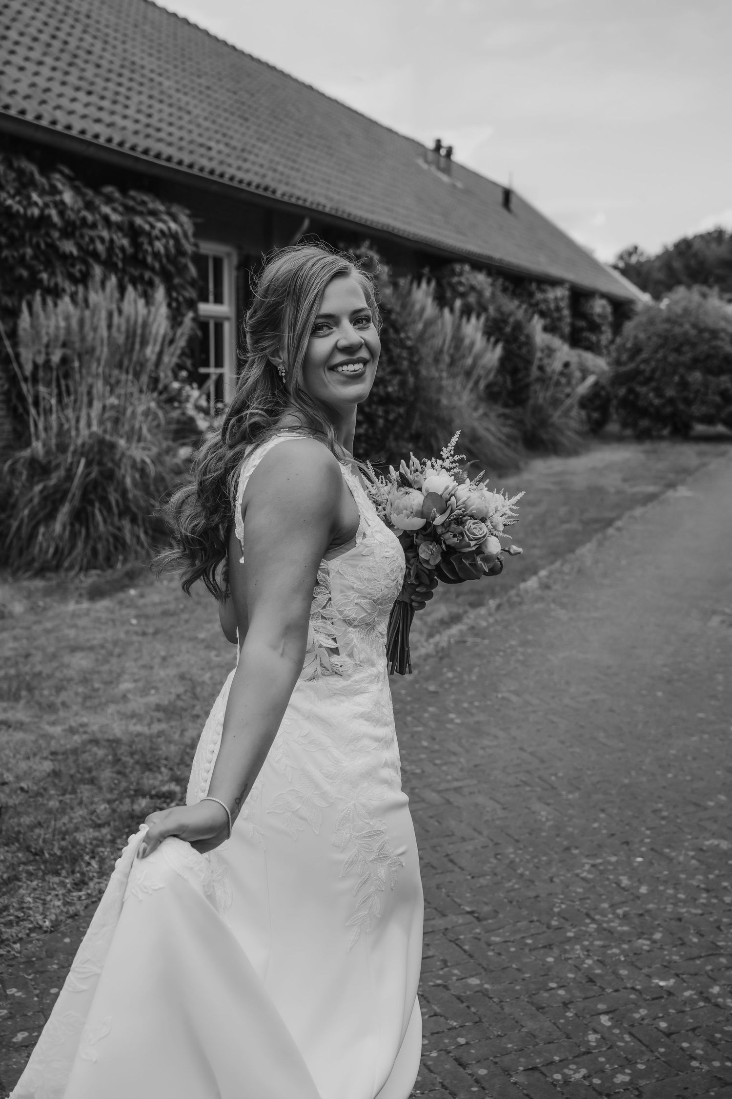 Een bruid in een witte trouwjurk loopt vrolijk over een tuinpad met een bruidsboeket in haar hand.