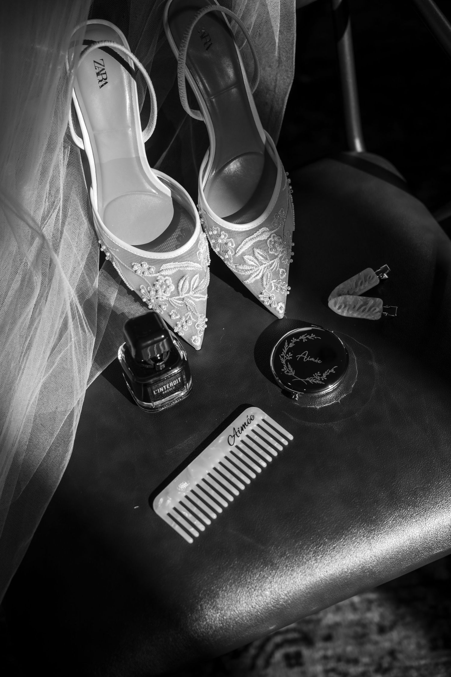 Black and white photo of elegant lace bridal heels beside perfume, compact, and comb on a dark surface.