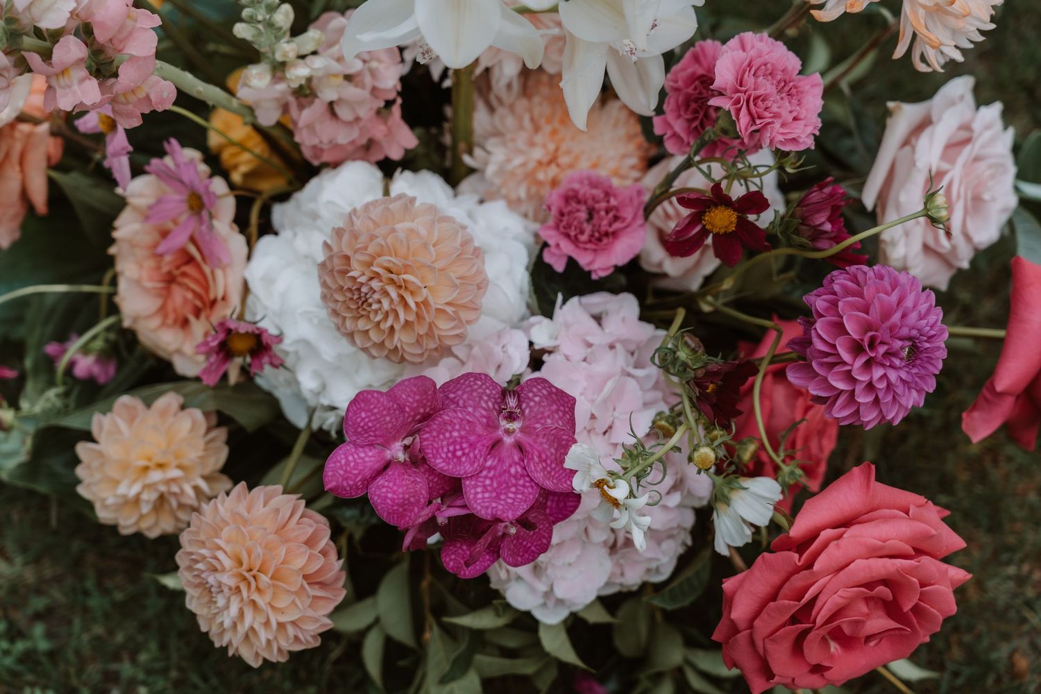 Beautiful floral arrangement featuring pink and peach dahlias, roses, and carnations in various shades against dark background.