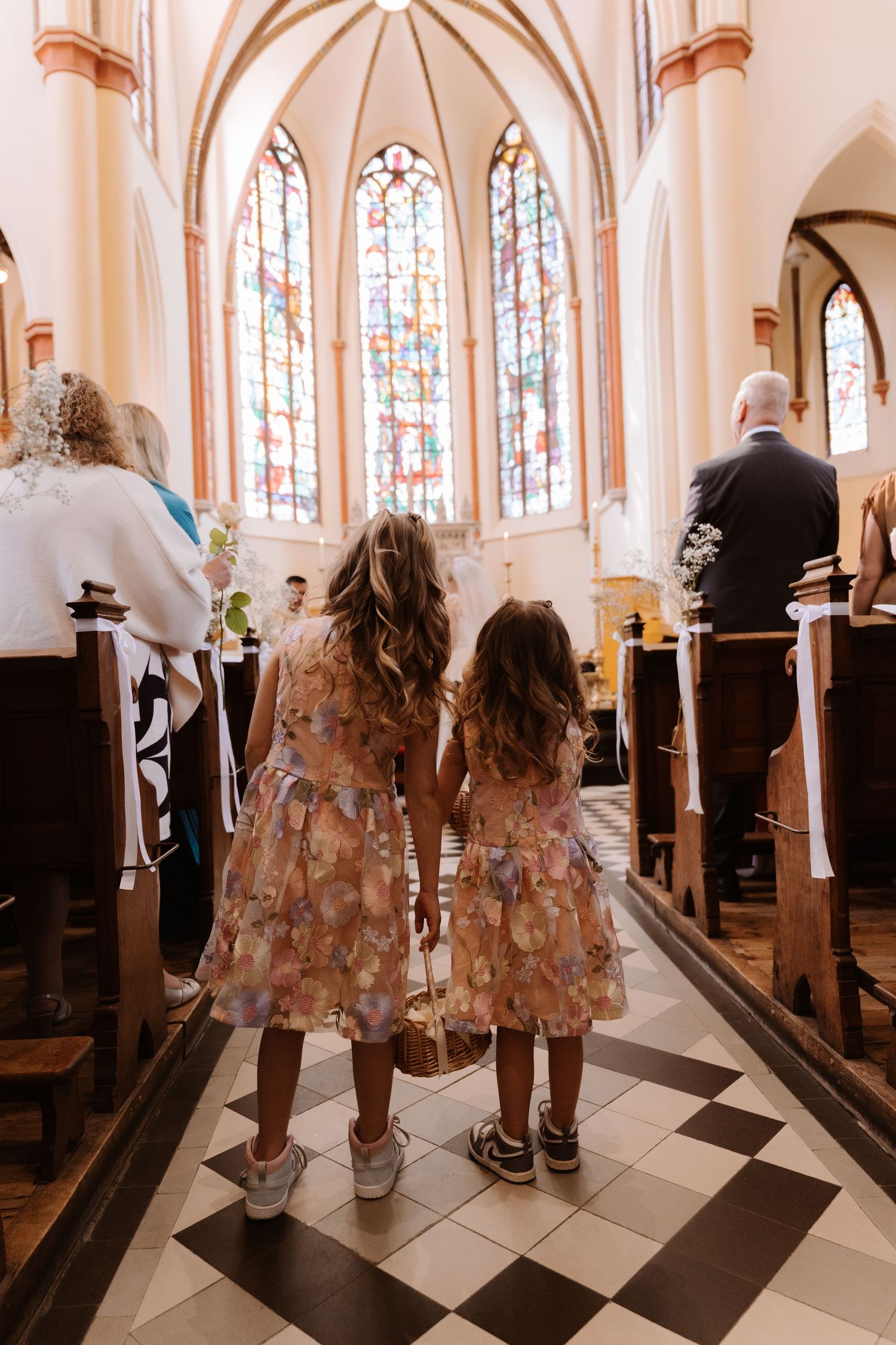 Two young girls in matching floral dresses hold hands walking down a church aisle during a wedding ceremony.