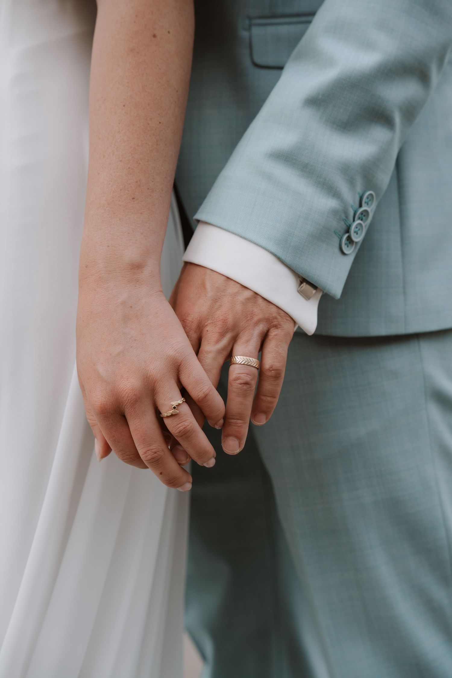 Close-up of couple's hands with wedding rings against light blue suit.