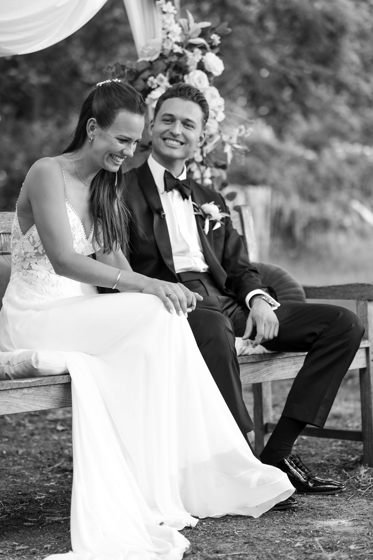A couple shares laughter while sitting together on a wooden bench during their wedding.