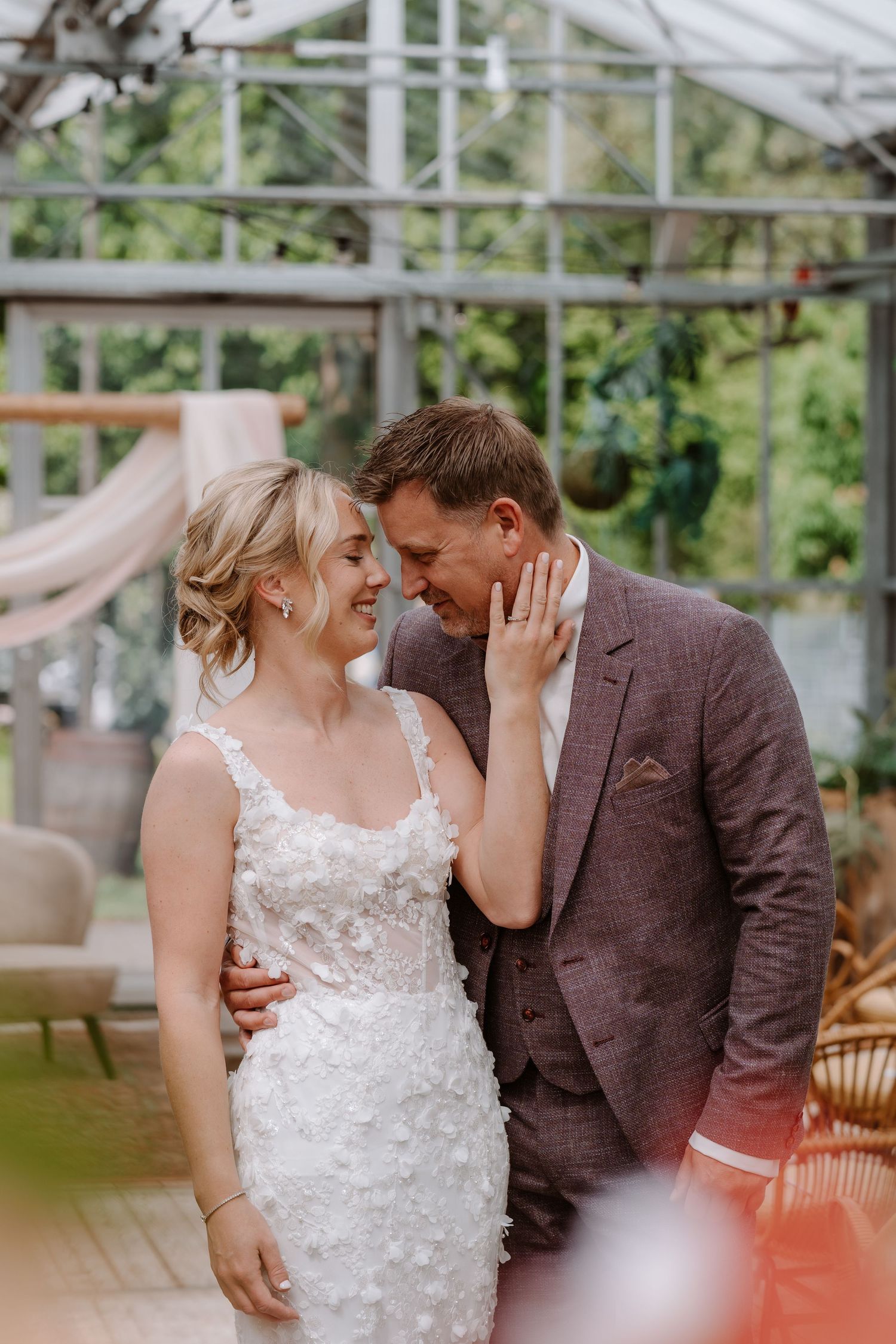 Bride and groom share a romantic moment in a greenhouse venue, bride in floral lace gown, groom in brown suit.