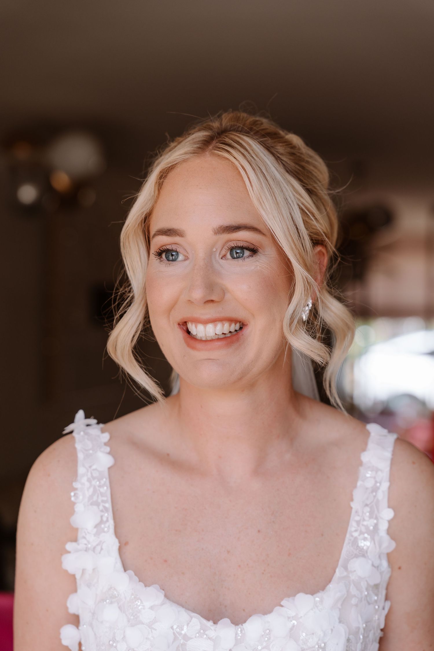 Smiling blonde bride in a white floral wedding dress with delicate drop earrings, captured in a bright indoor setting.