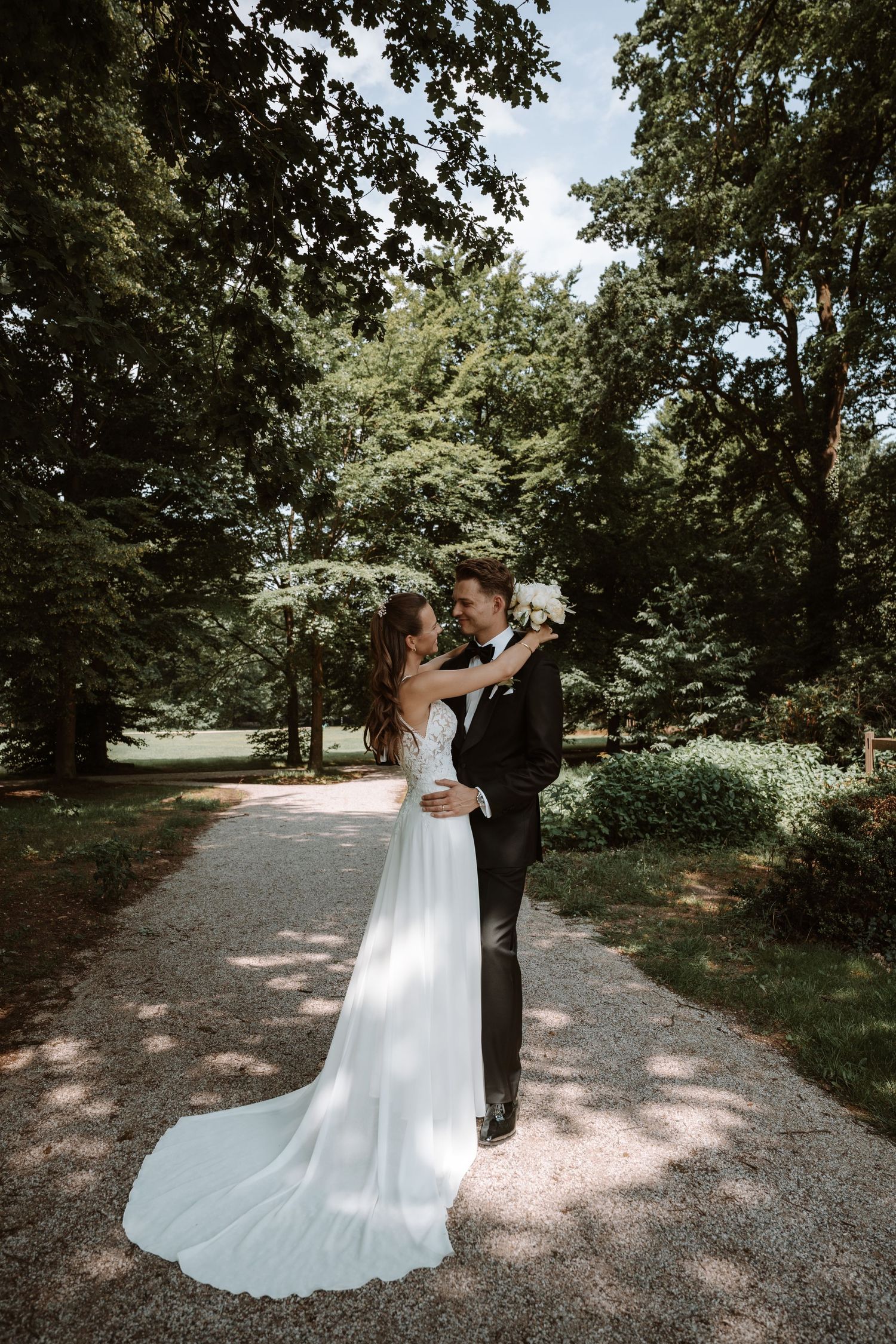 A romantic wedding couple embraces on a tree-lined gravel path in a garden setting.