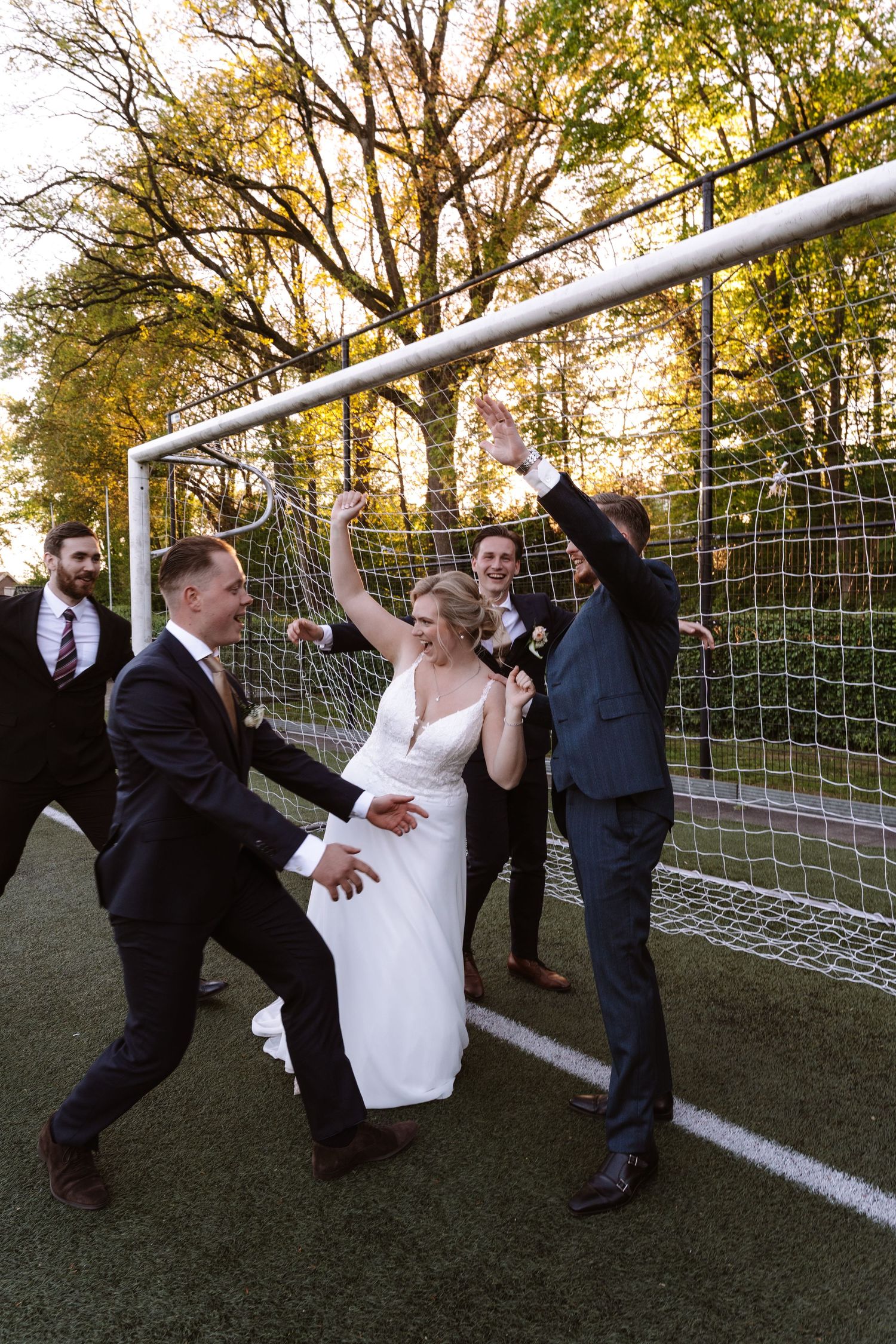 Bride and groom celebrate with wedding party on a soccer field in front of a goal, surrounded by lush autumn trees.