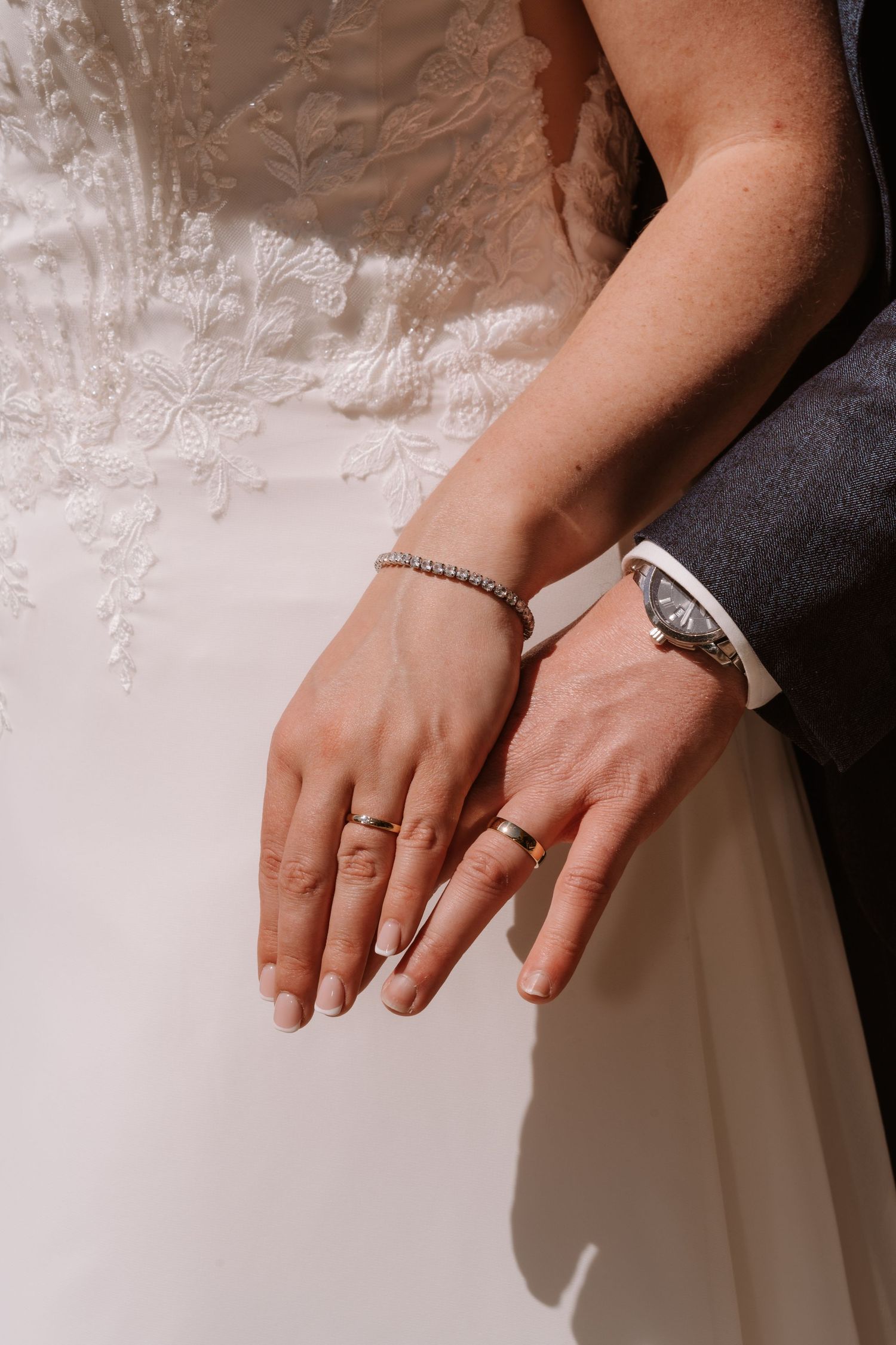 Bride and groom displaying wedding rings, bride wearing diamond bracelet, groom in gray suit with watch.