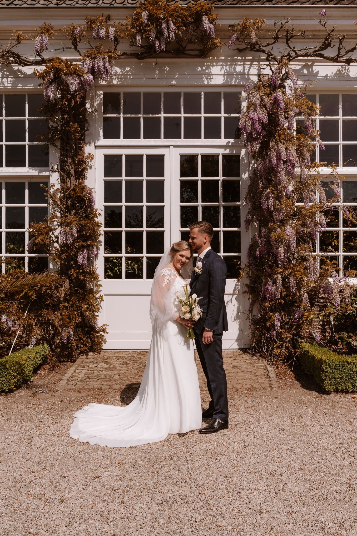Bride and groom stand together before a wisteria-covered building on their wedding day.