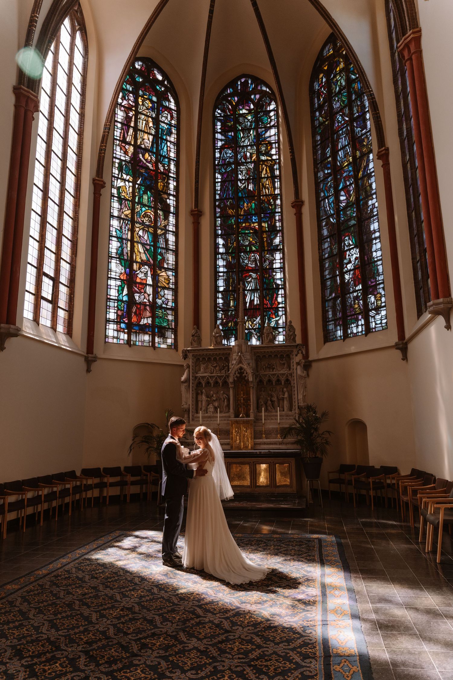 Bride and groom embrace before stained glass windows at a church altar, bathed in warm natural light.