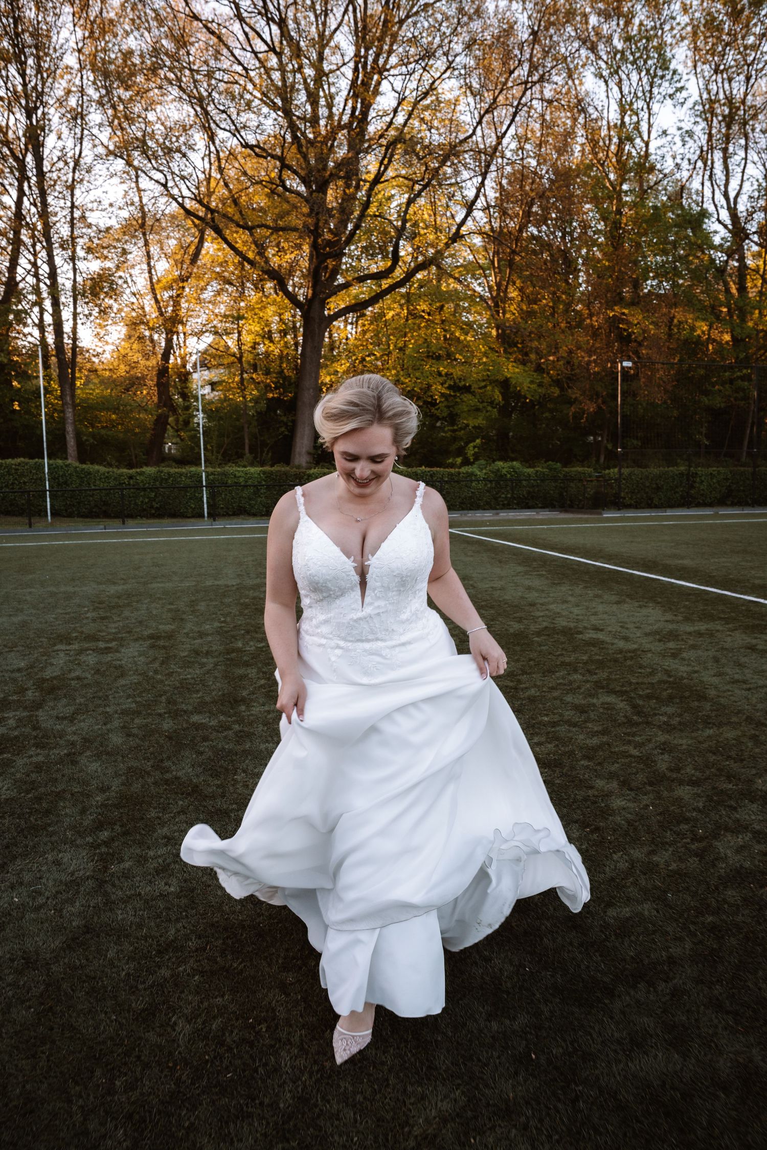 Bride in white lace wedding gown twirling on green lawn with autumn trees in golden light.