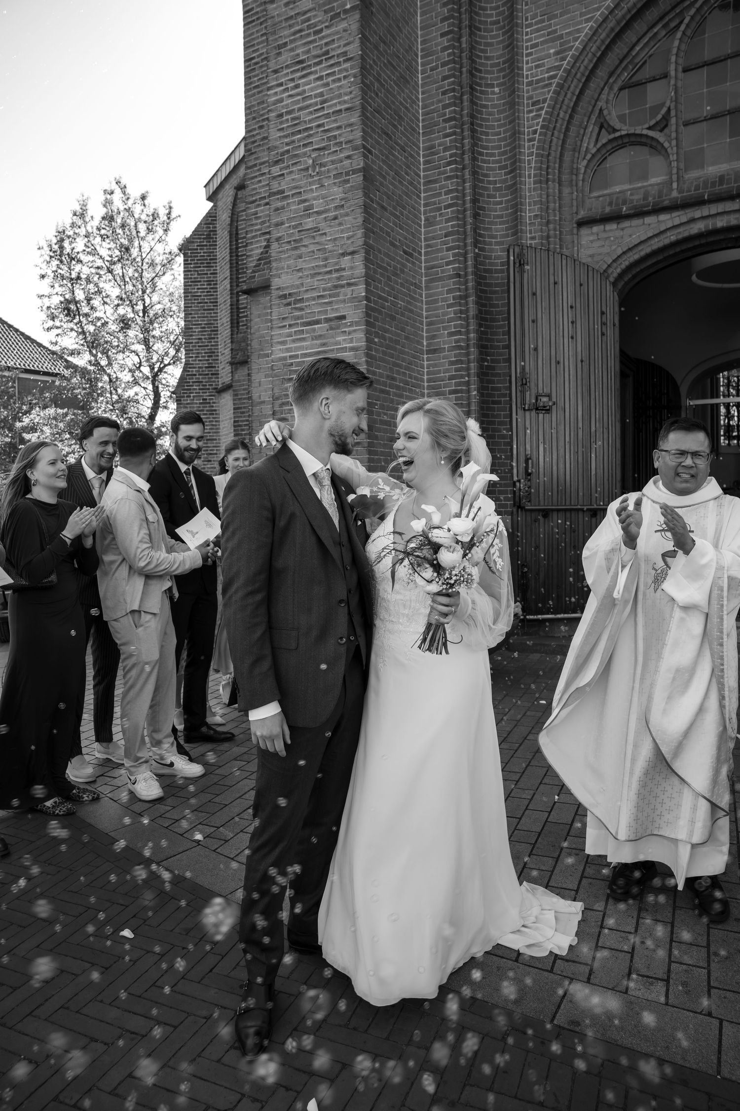 Black and white photo of a newlywed couple celebrating outside a brick church with confetti and guests clapping.