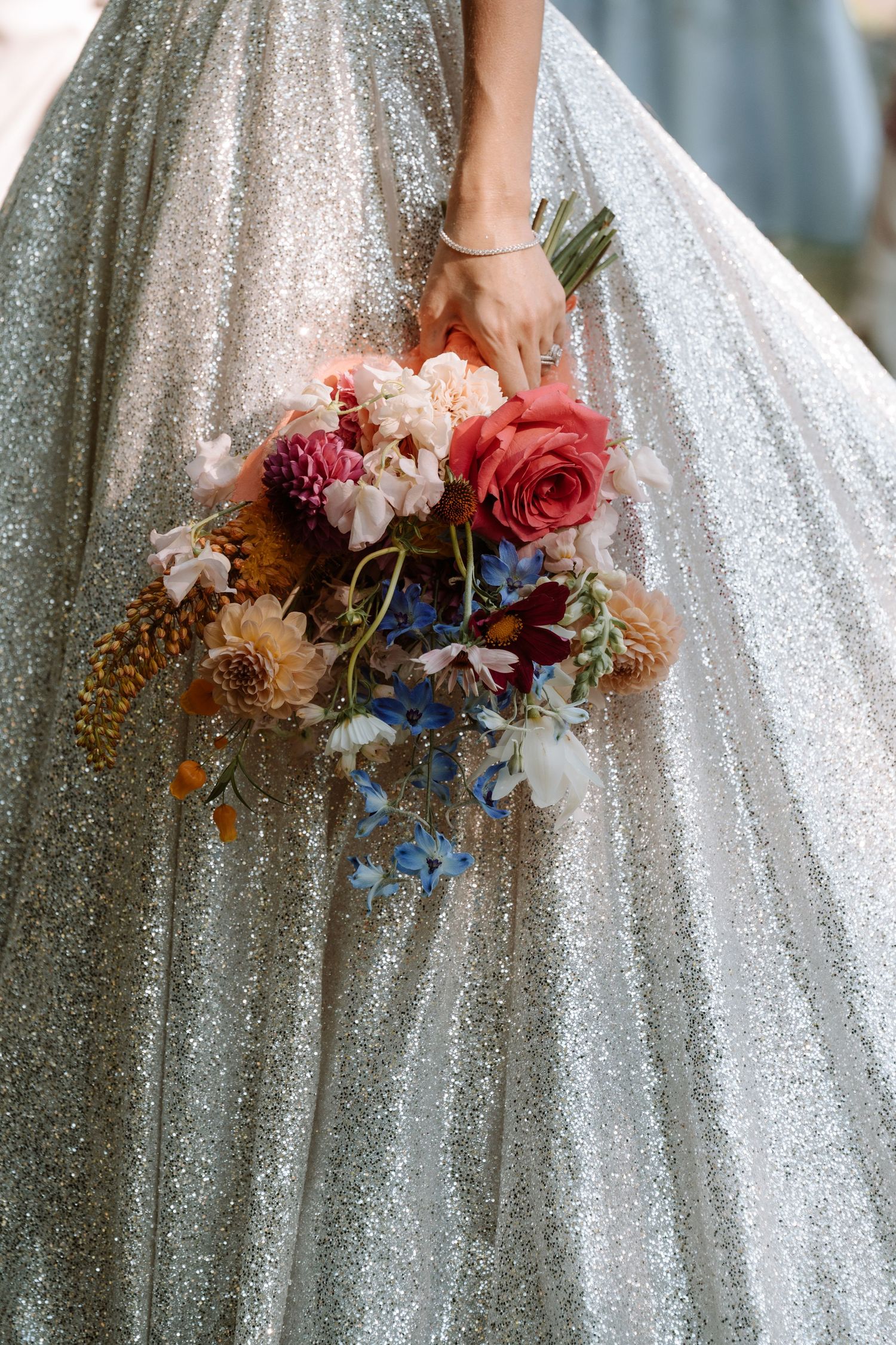 Close-up view of a sparkly white wedding gown with a colorful bouquet featuring pink roses and blue flowers.