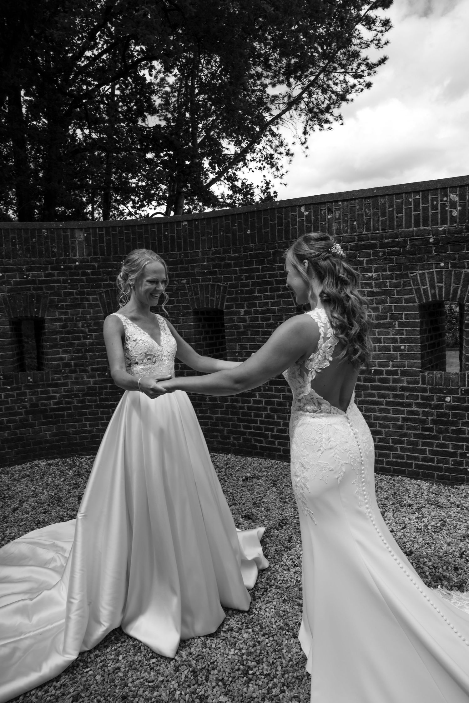 Two people in elegant white wedding gowns standing together against brick wall.