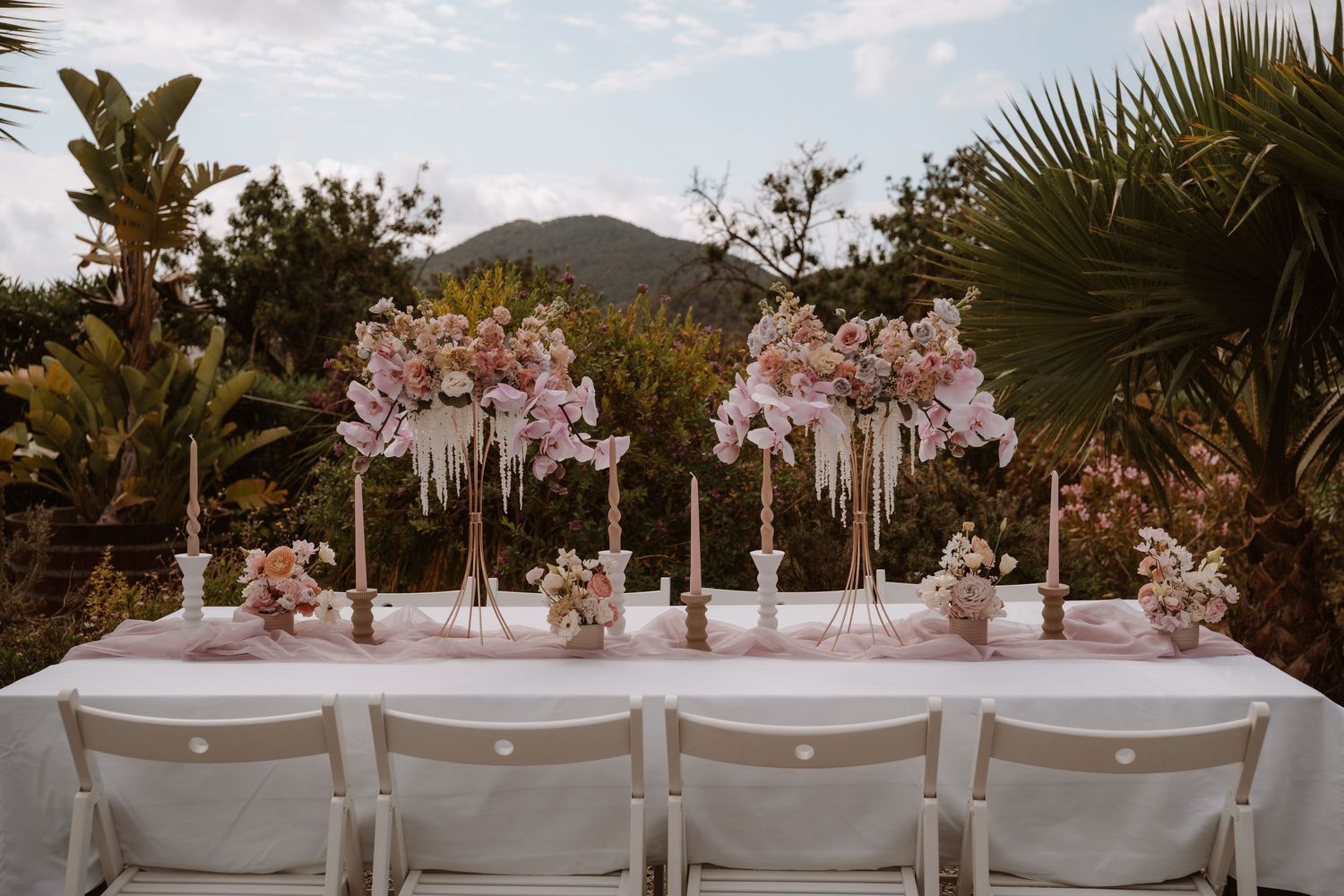 Elegant outdoor wedding reception table setup with pink floral arrangements and palm trees in the background.