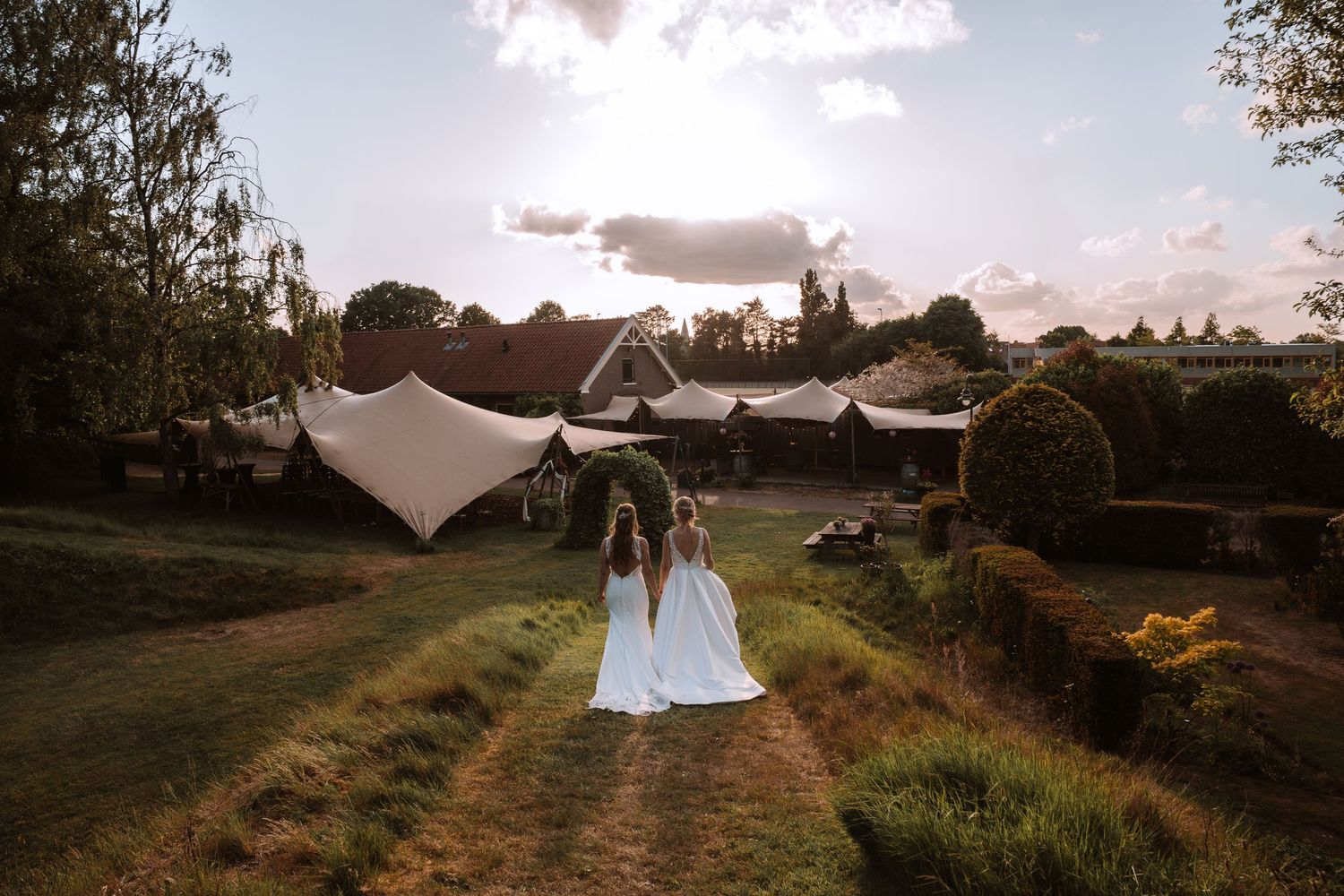 A couple walks down a path at sunset with white tent canopies in the background at an outdoor wedding venue.