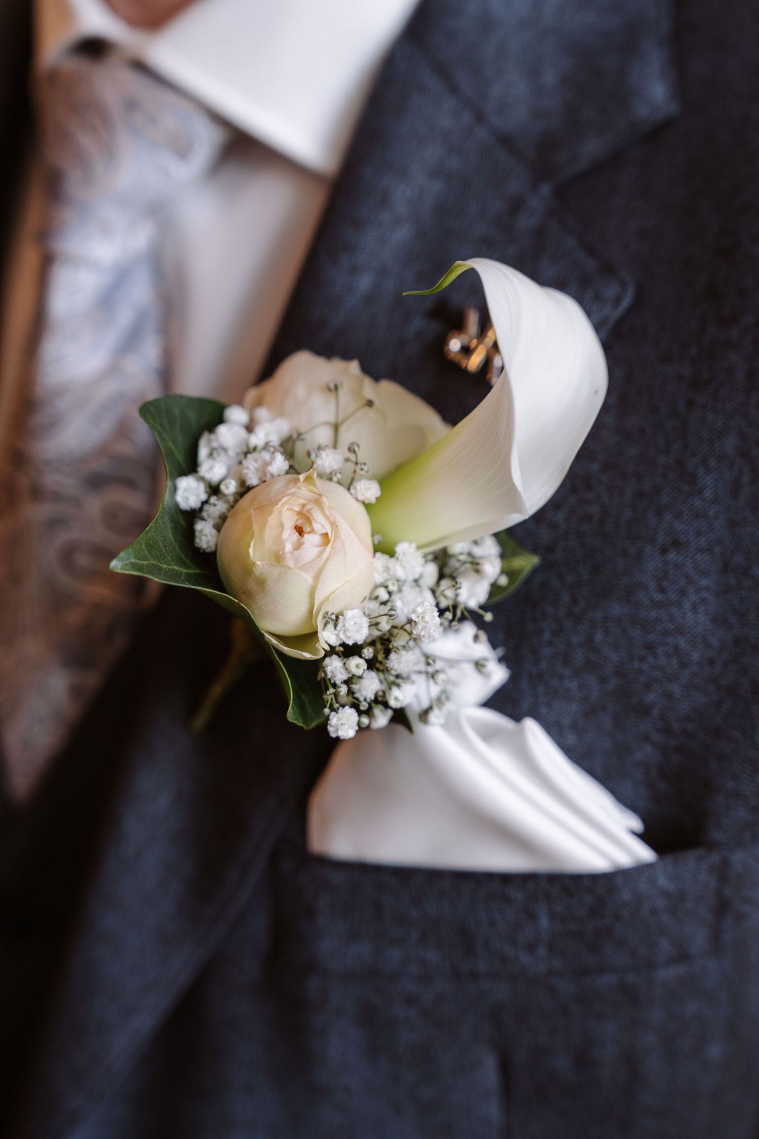 Close-up of a wedding boutonniere with white calla lily, cream rose, and baby's breath on a gray suit lapel.