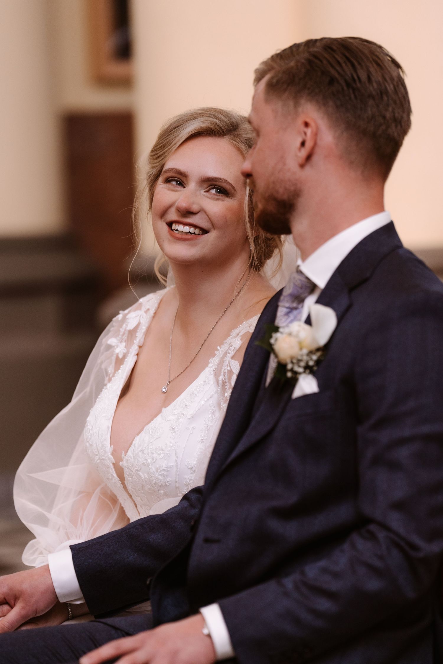 Smiling bride in lace wedding dress gazes at groom in navy suit with boutonniere during wedding ceremony.