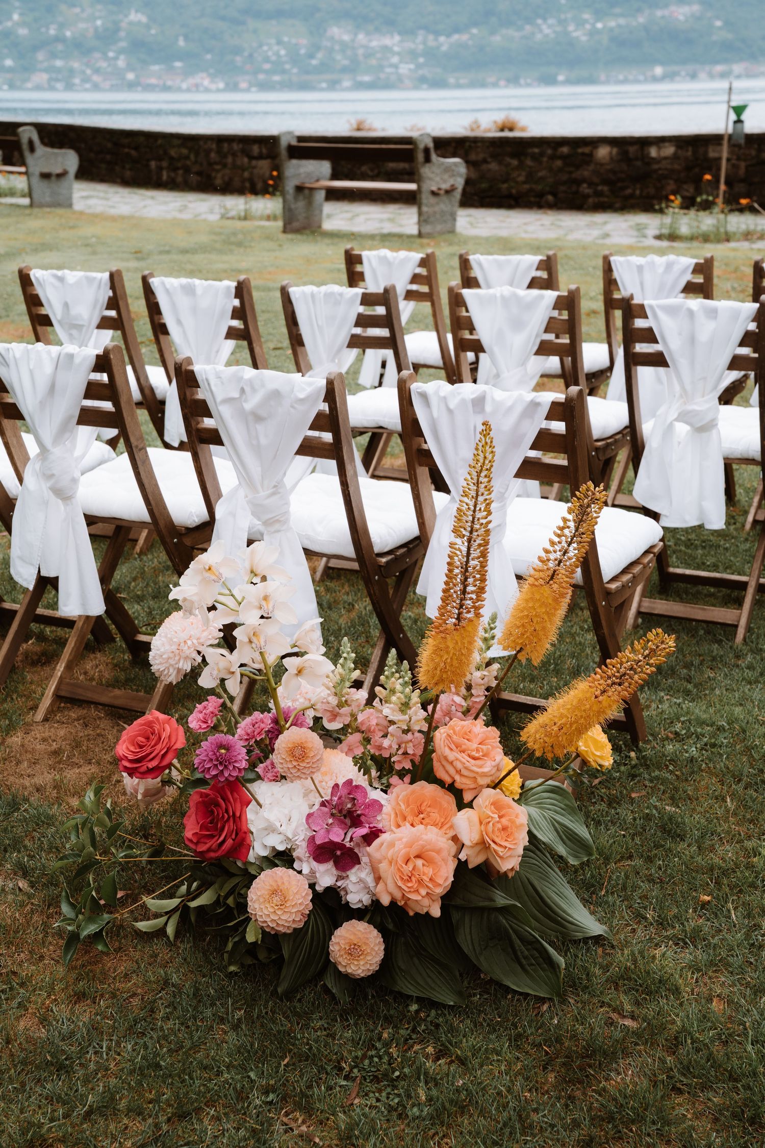 Rustic wooden chairs decorated with white fabric bows and colorful floral arrangements for an outdoor wedding ceremony.