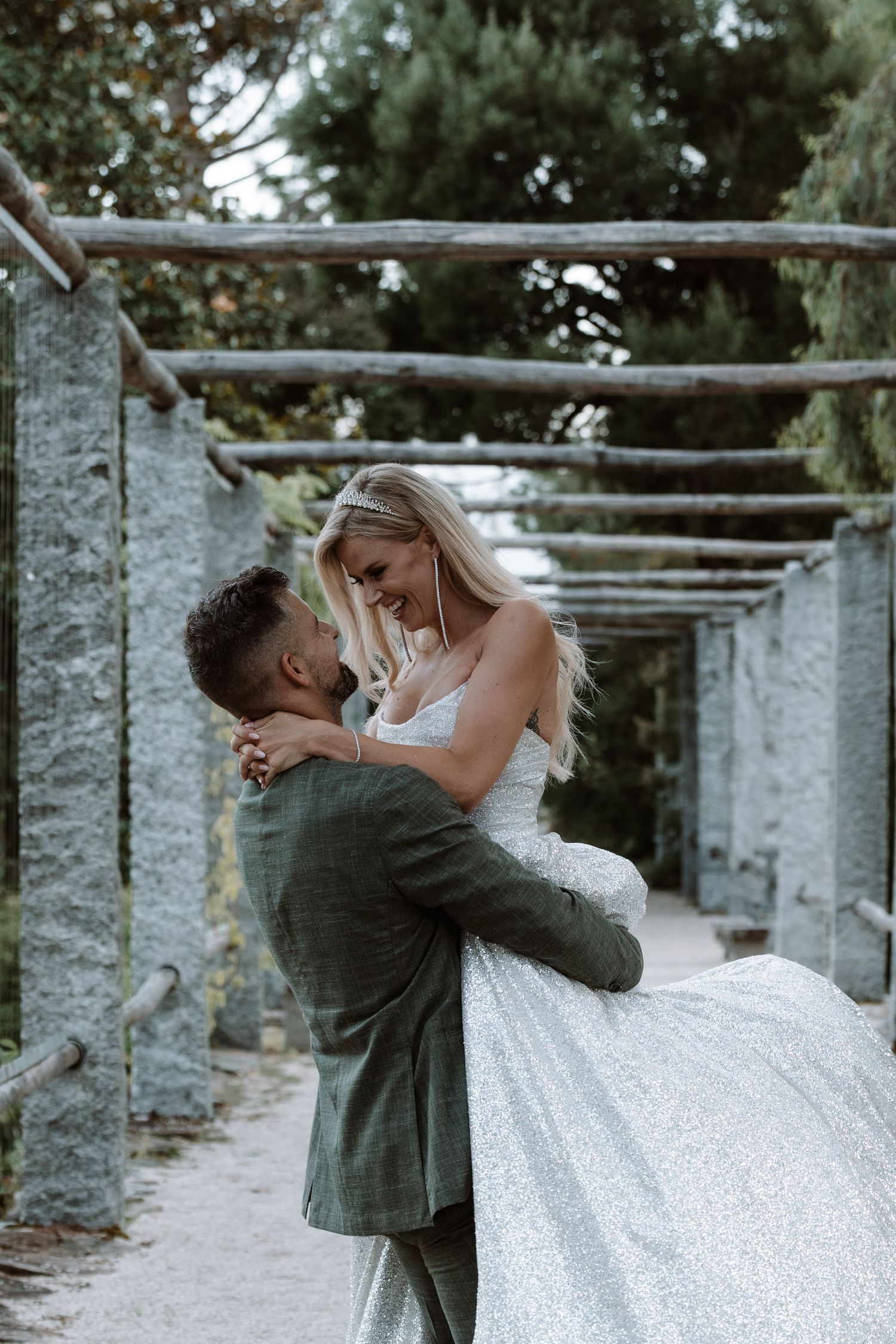A romantic wedding couple embraces under a rustic wooden pergola in an outdoor garden setting.