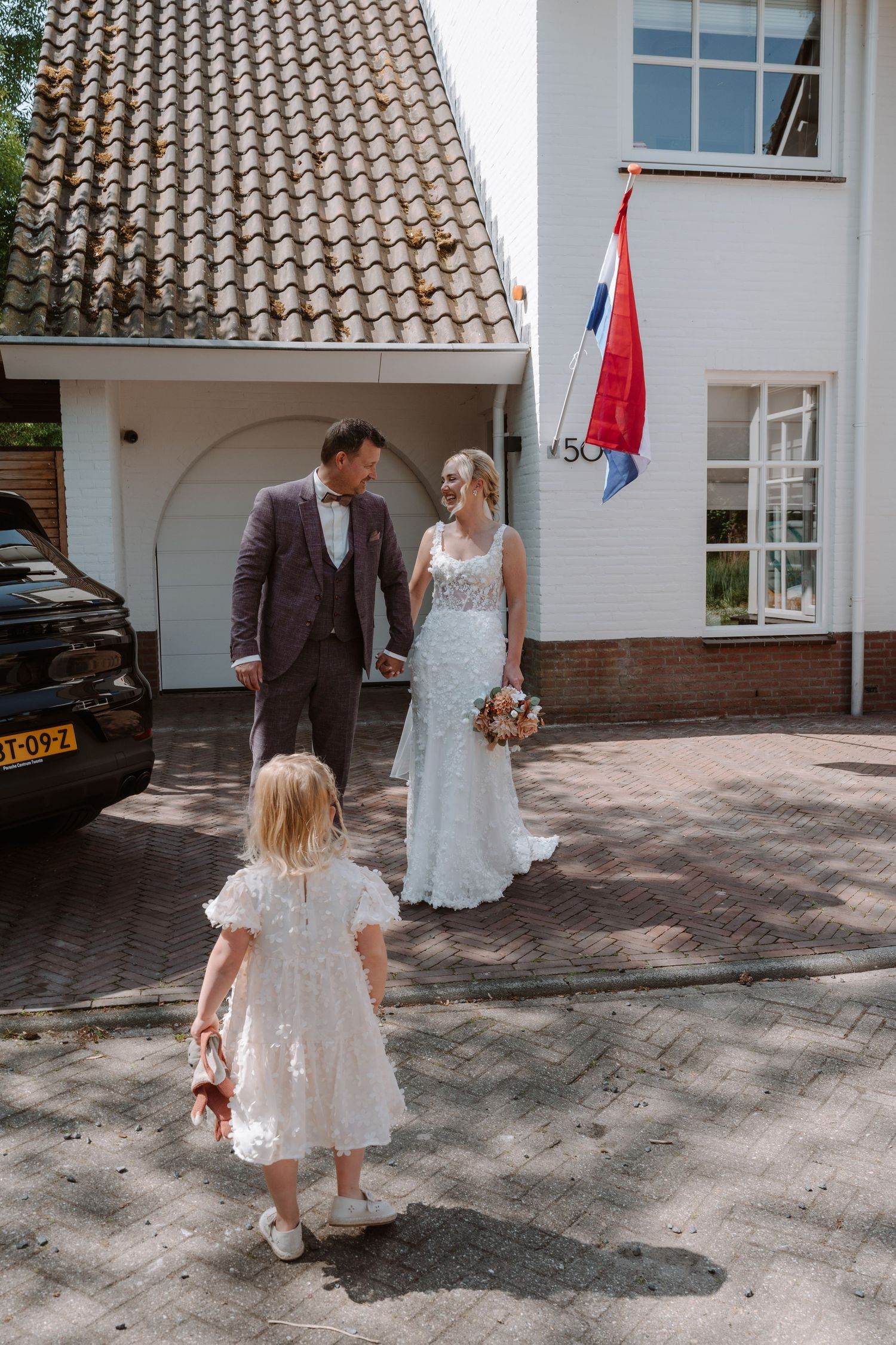Bride and groom walking with flower girl outside white Dutch house with Netherlands flag on sunny wedding day.