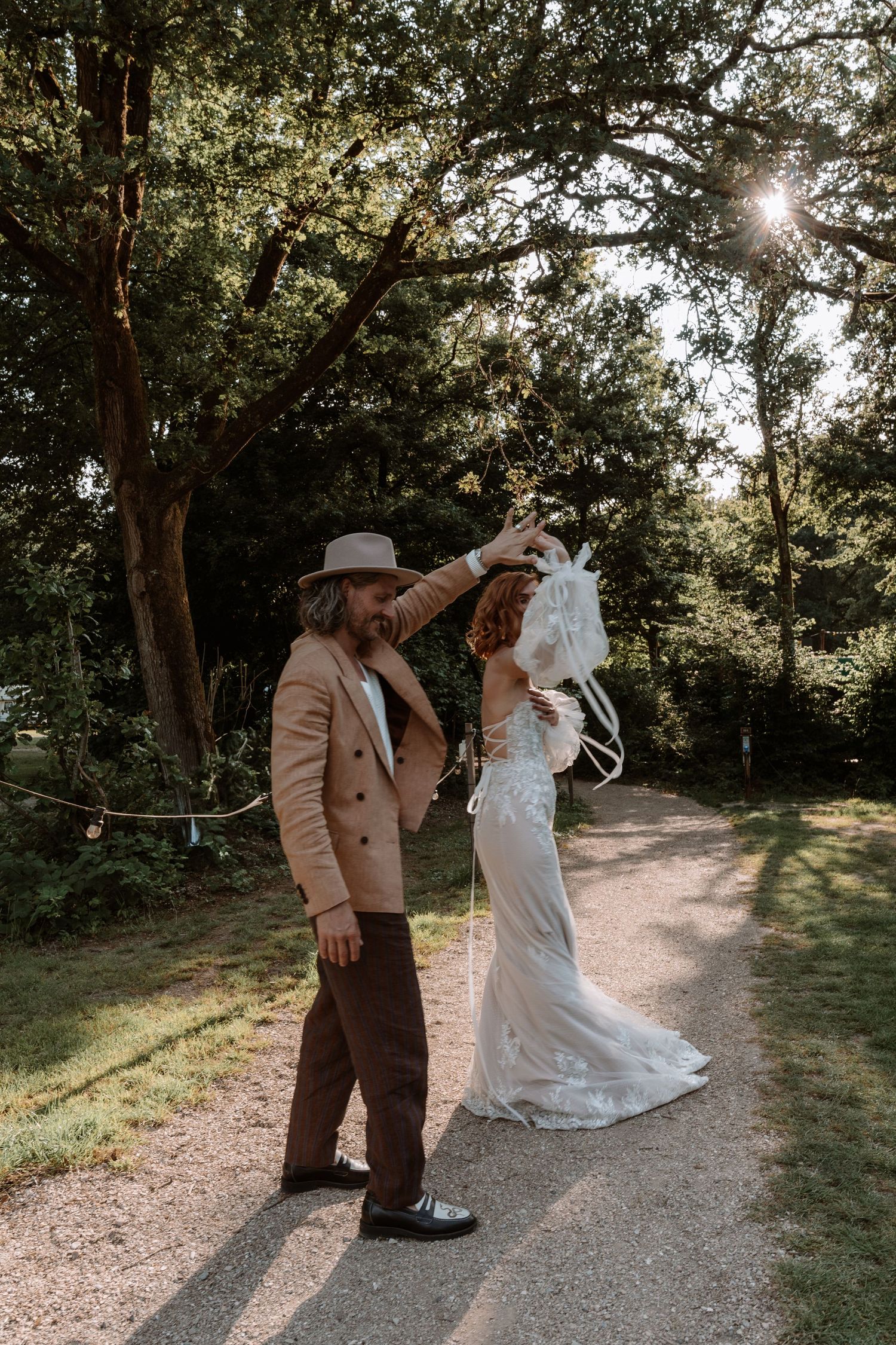 A couple dances and twirls on a sunlit forest path in their wedding attire.