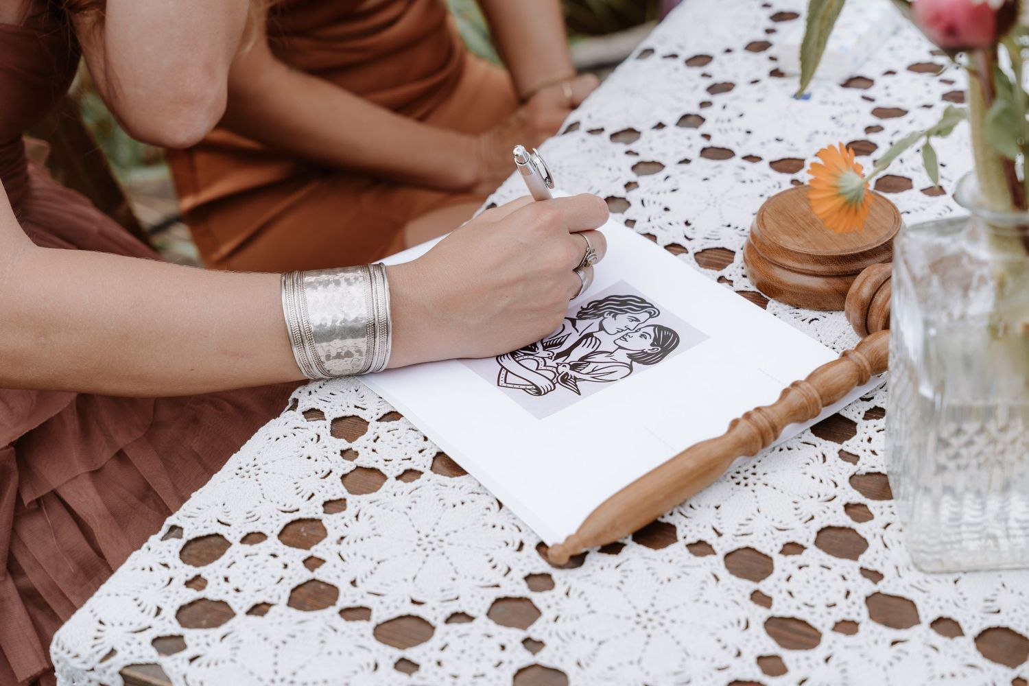 Hand wearing silver bracelet sketching on white lace tablecloth during wedding celebration.
