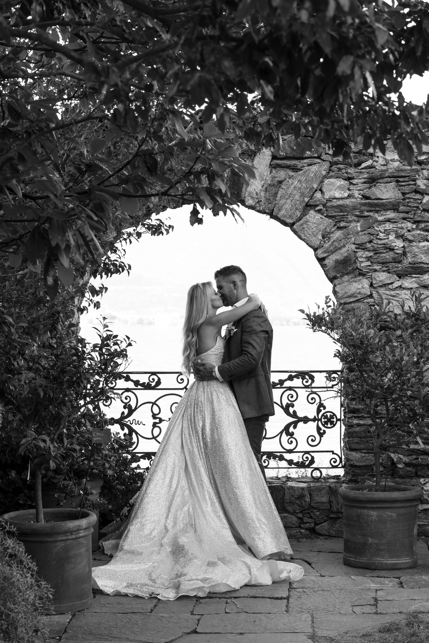 A romantic black and white wedding photo showing a couple embracing under a stone archway with ornate iron railings.