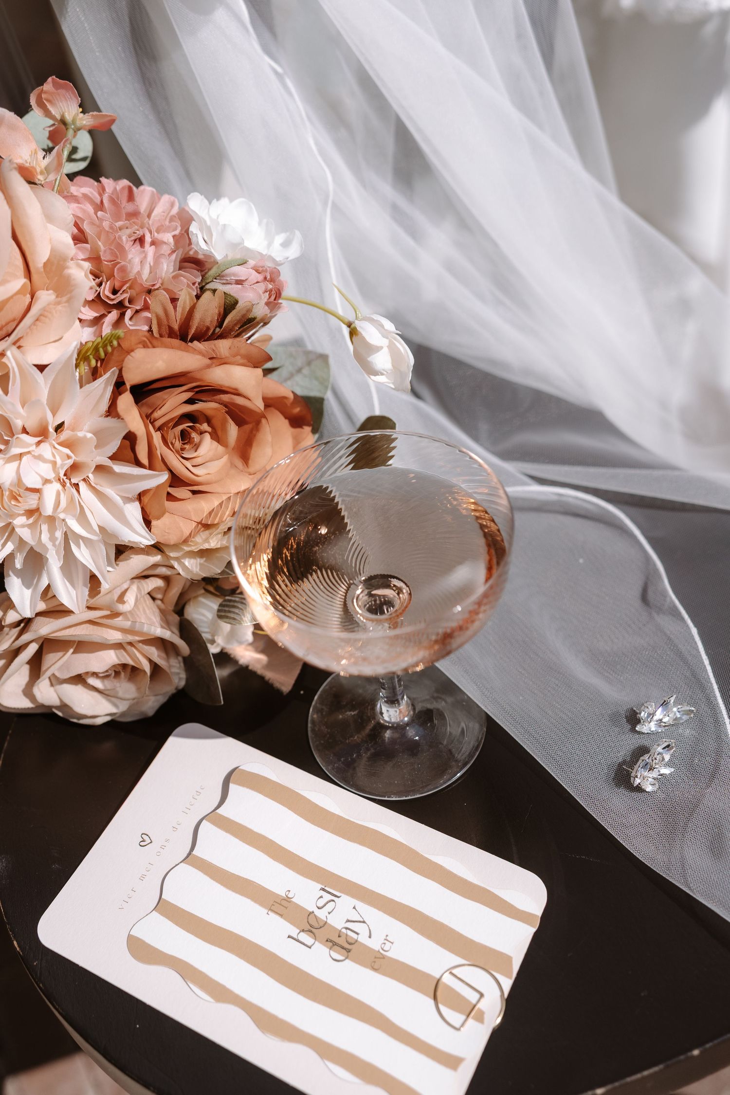 Elegant wedding flat lay with blush roses, champagne coupe glass, diamond earrings, and striped card on dark table.
