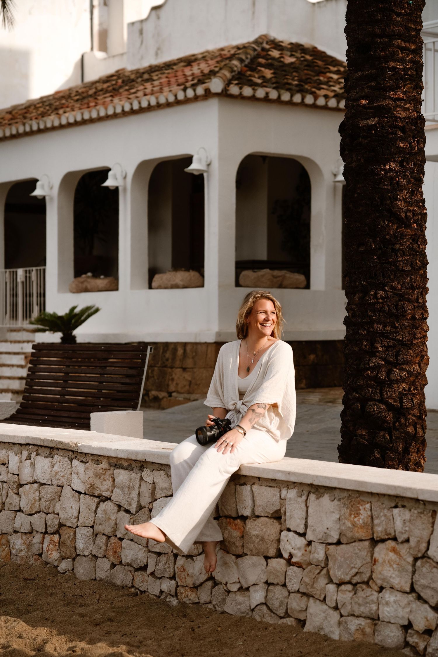 Photographer in white dress sits on stone wall near Mediterranean-style building with palm trees.