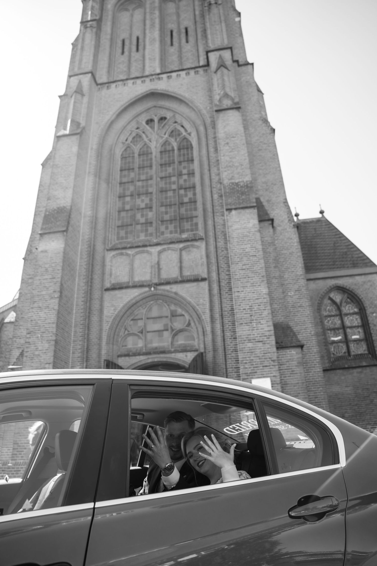 Black and white photo of a couple kissing in a car outside a historic church tower.