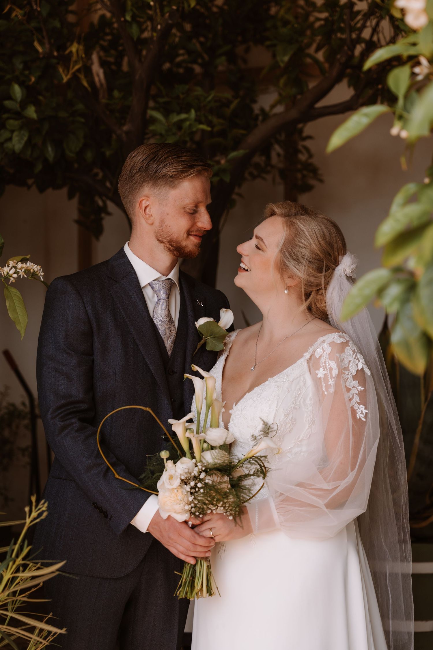 A smiling bride in a lace wedding dress holding a bouquet stands with a groom in a dark suit surrounded by greenery.