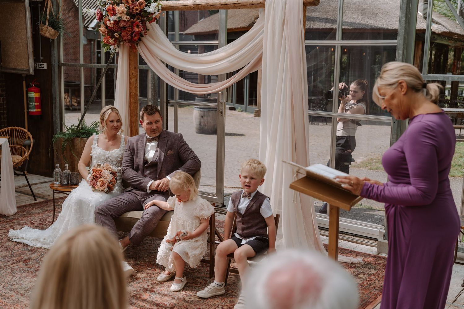 Wedding ceremony with bride, groom, and children seated under floral arch as officiant in purple dress reads vows.