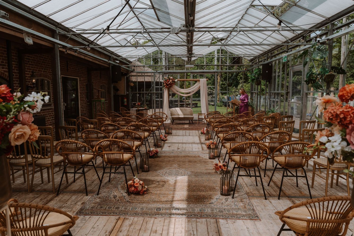 Elegant greenhouse wedding ceremony setup with rattan chairs, floral arrangements, and a draped arch in a sunlit glass venue.
