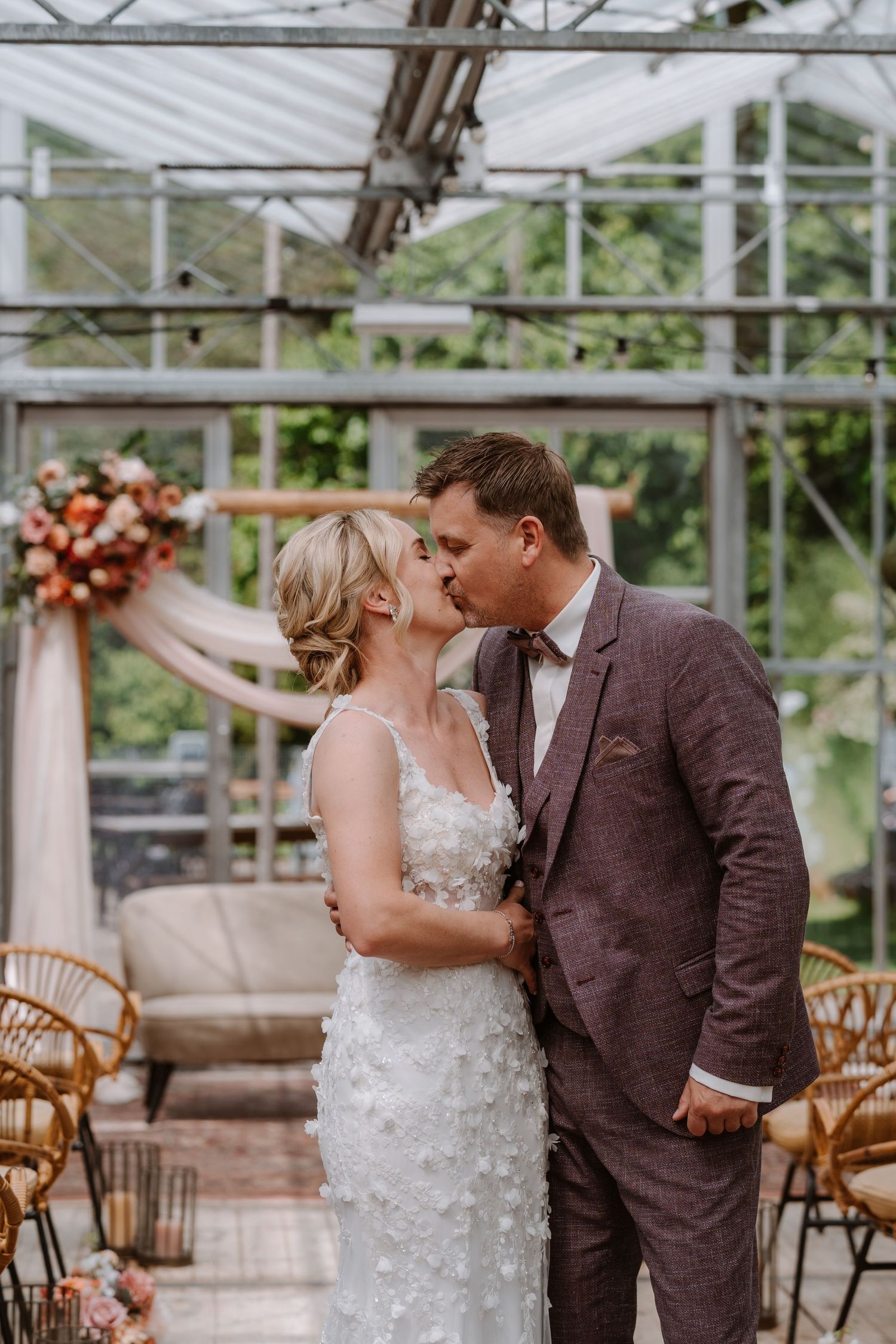 Bride and groom share first kiss at greenhouse wedding ceremony with floral arch and rattan chairs.