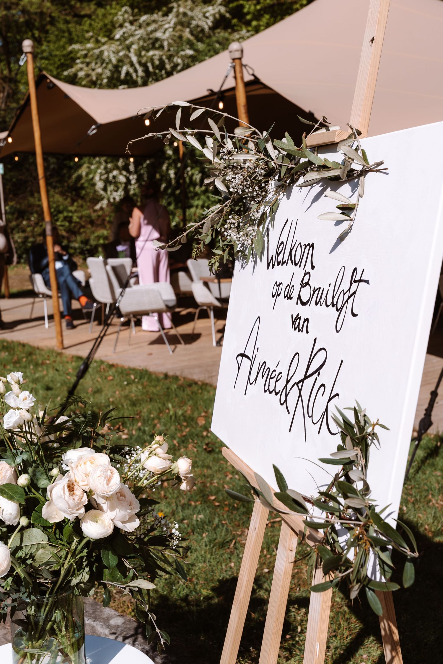 Elegant outdoor wedding welcome sign on easel with floral greenery decor, reading Welkom op de Bruiloft van Aimee & Rick.