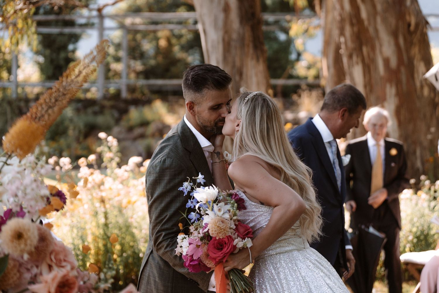 Wedding ceremony guests gather outdoors under tall trees during a golden sunset celebration.