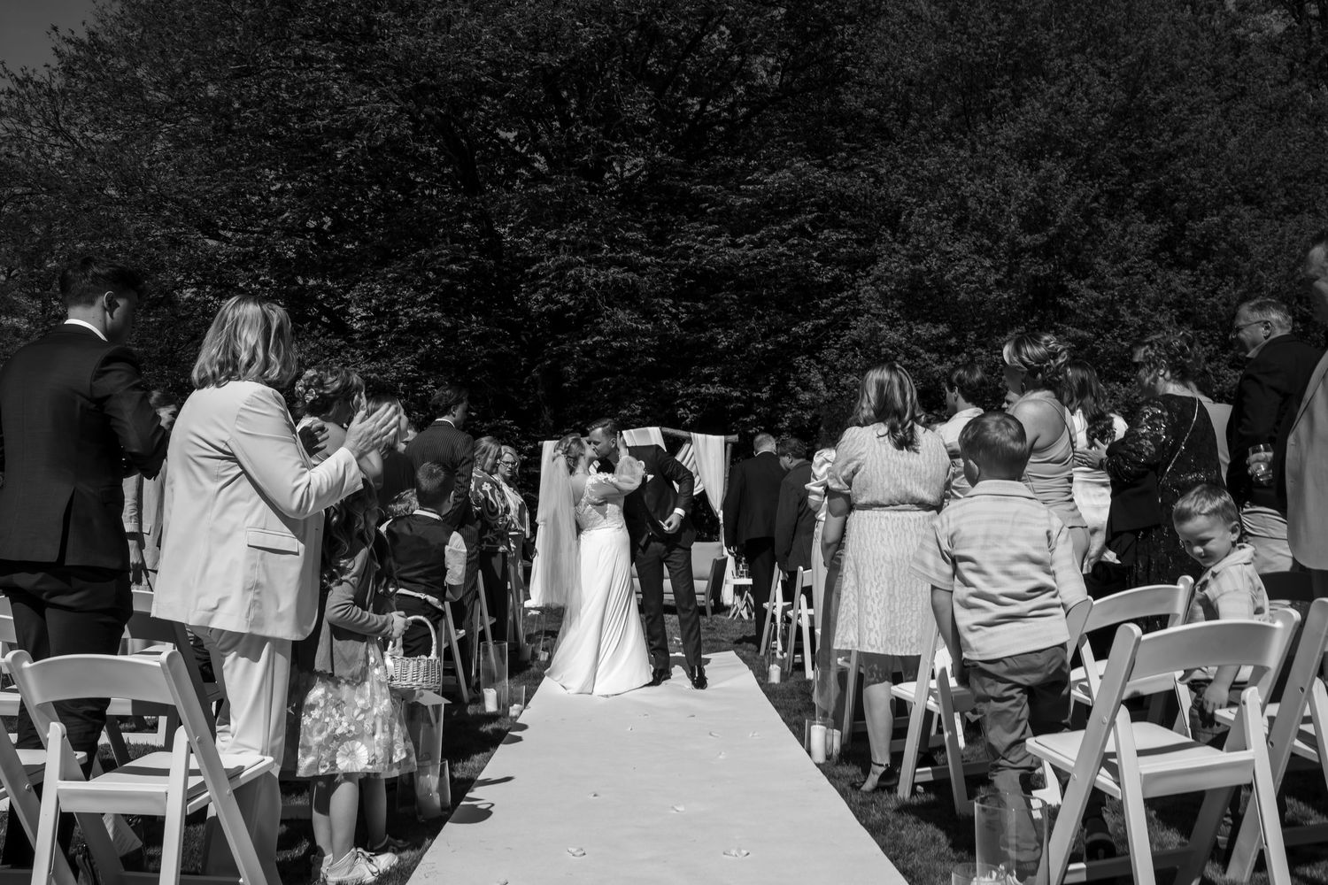 Black and white outdoor wedding ceremony with guests applauding as bride and groom walk down the aisle.