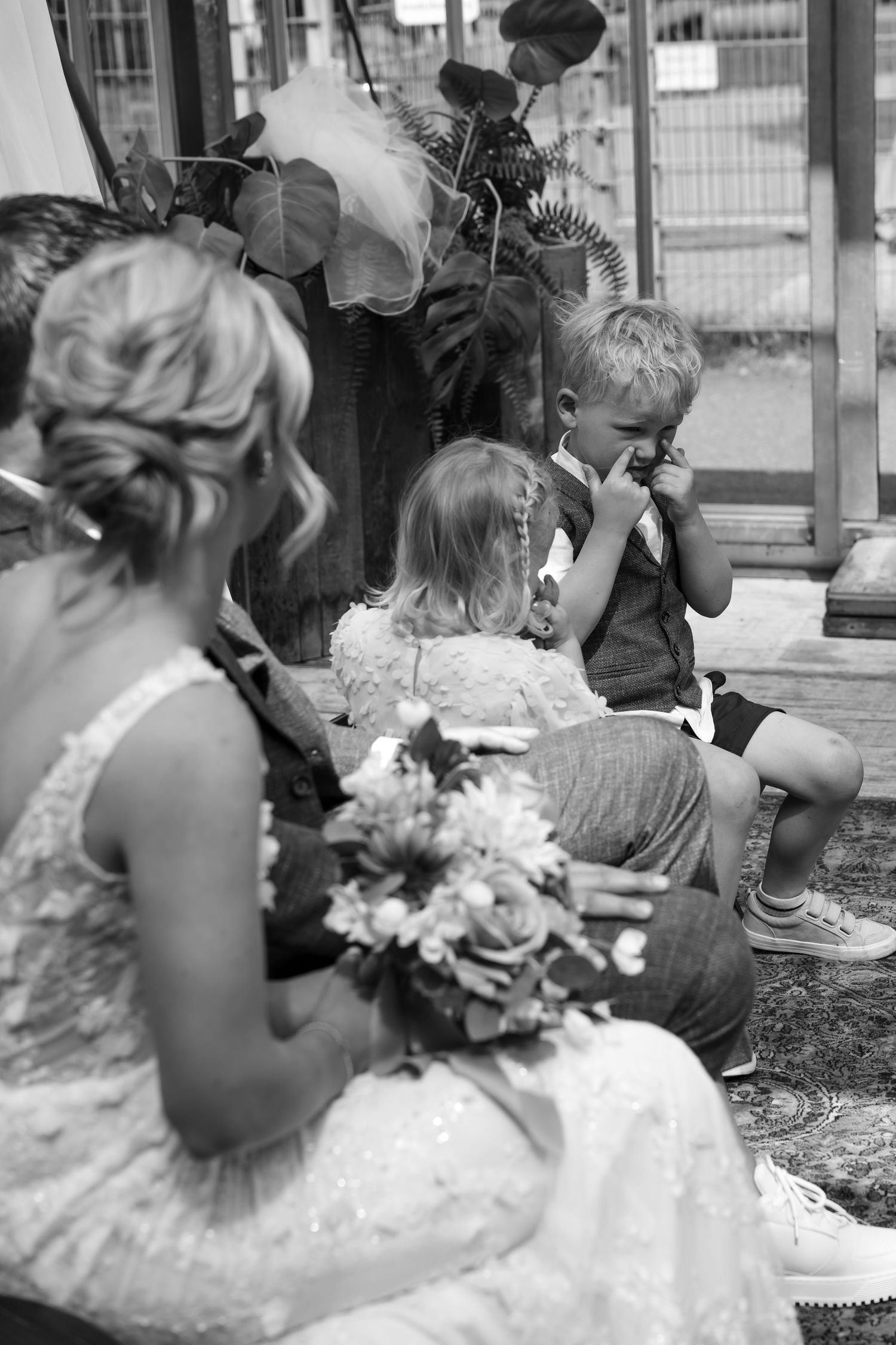 Black and white photo of a bride holding bouquet sitting with young children during a wedding ceremony outdoors.