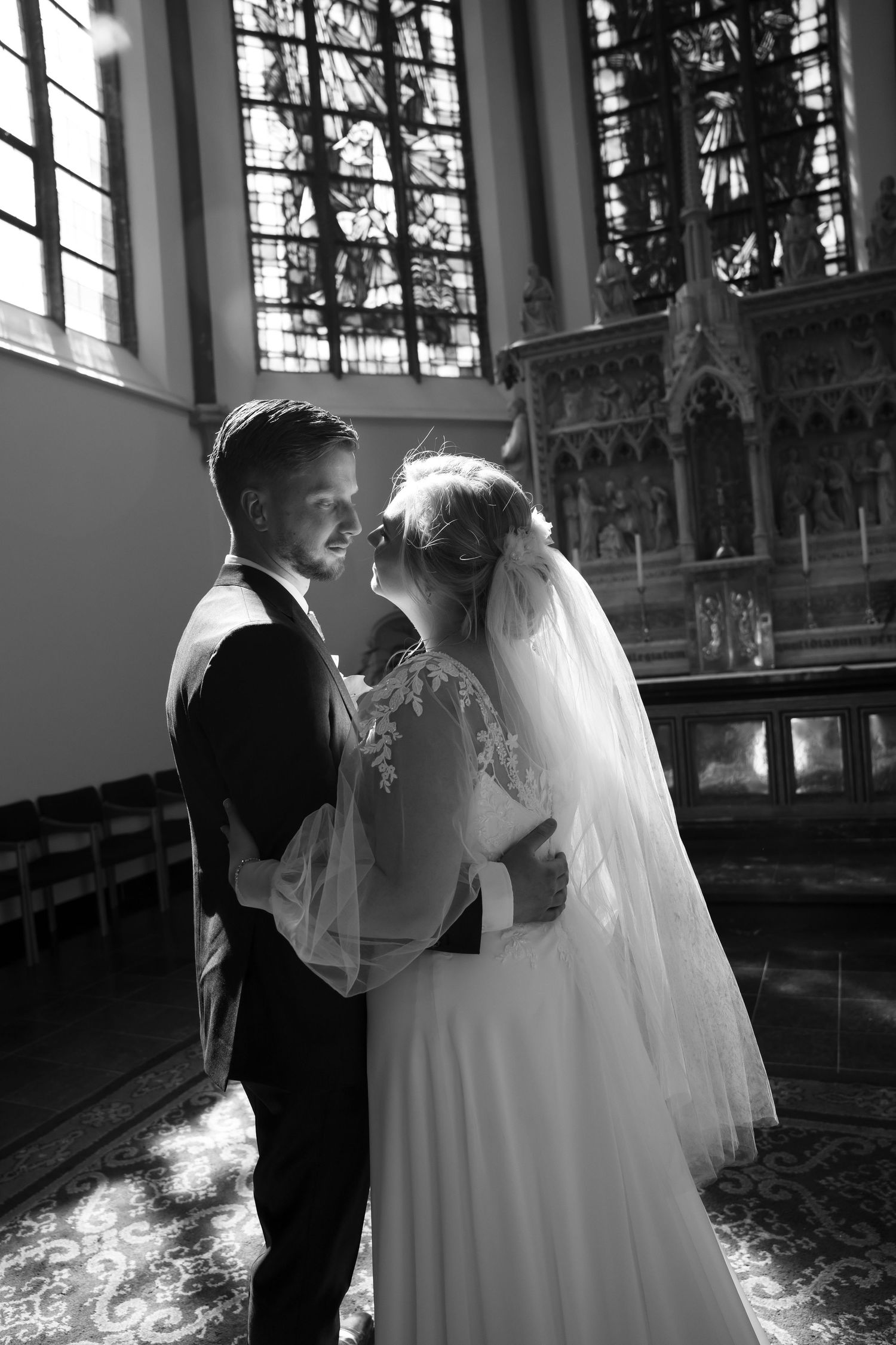 Black and white photo of a bride and groom sharing a tender moment inside a sunlit church with stained glass windows.