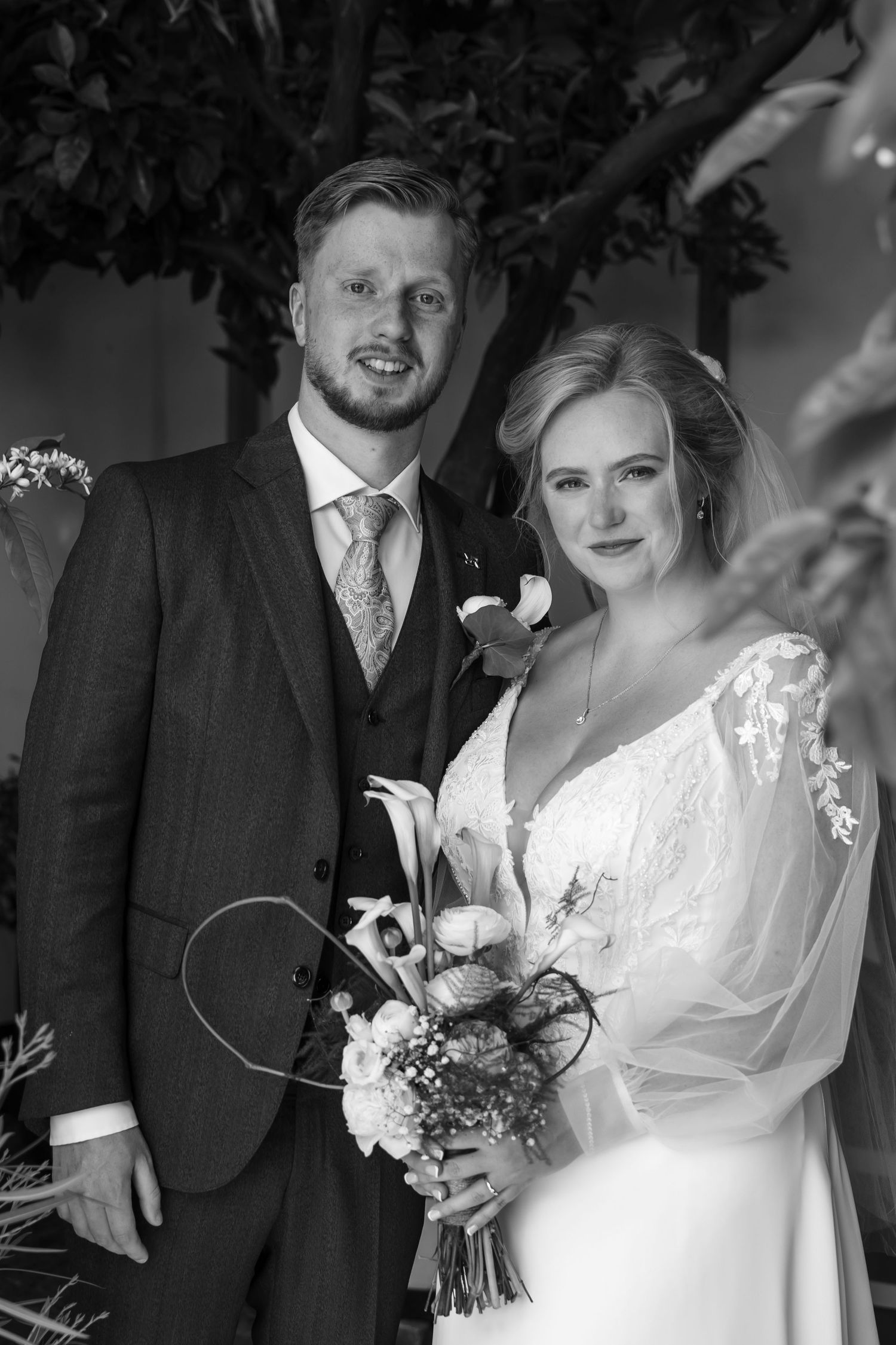 Black and white wedding portrait of a smiling bride holding a bouquet and groom in a suit posing together outdoors.