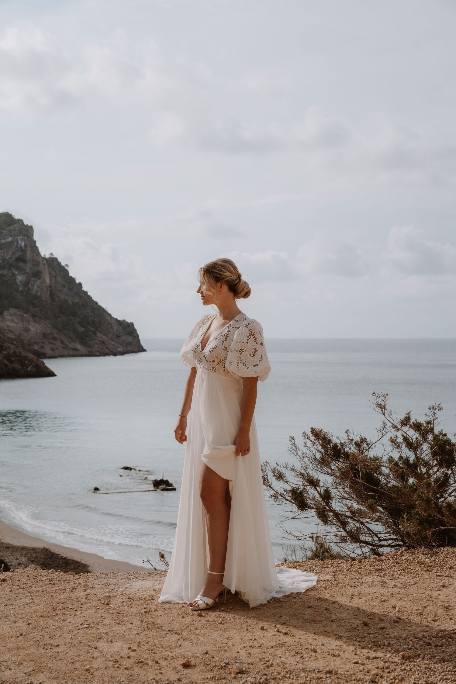 White wedding dress flows in coastal breeze against rocky beach backdrop.