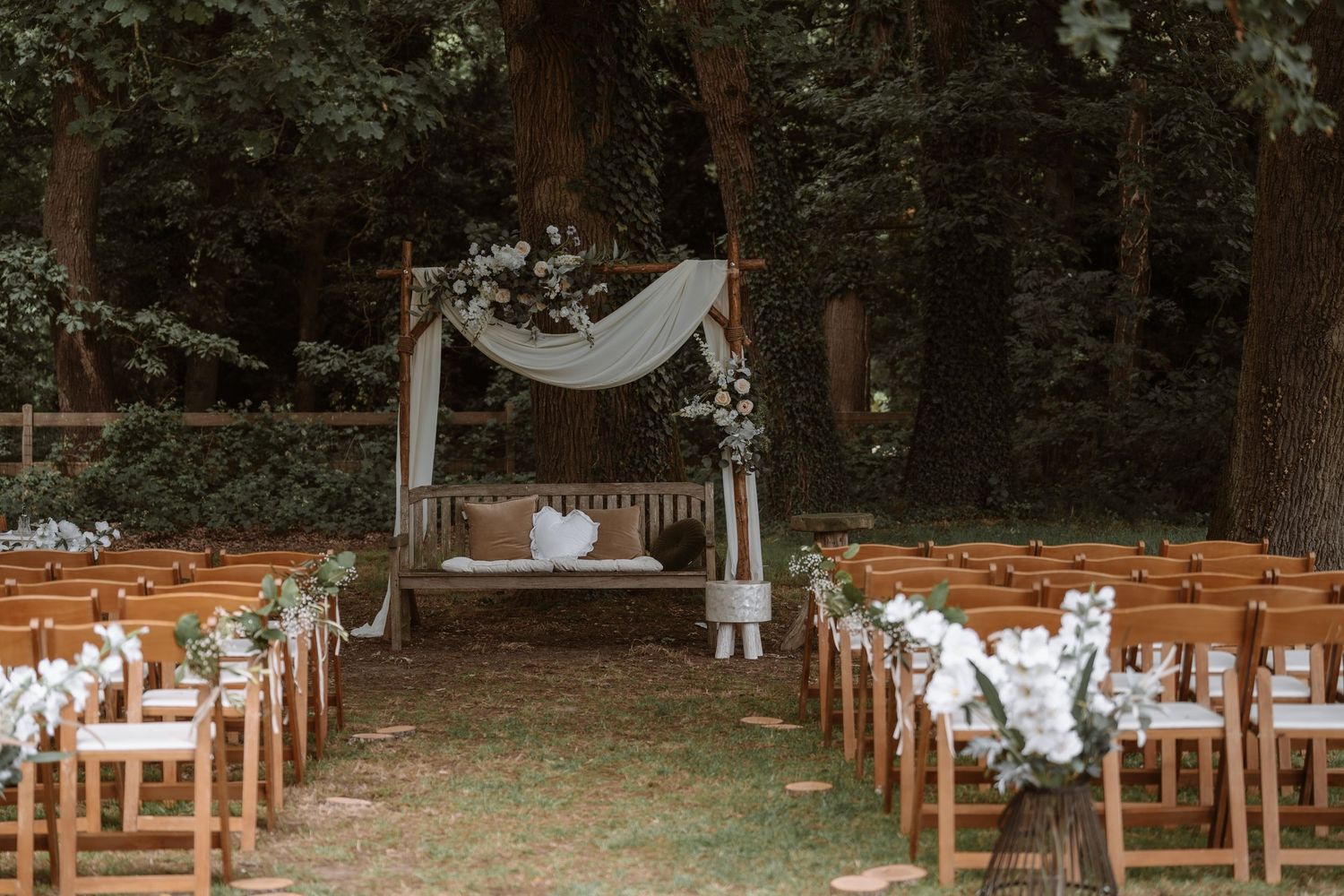 Rustic outdoor wedding ceremony setup with wooden chairs and floral decorations in a wooded setting.
