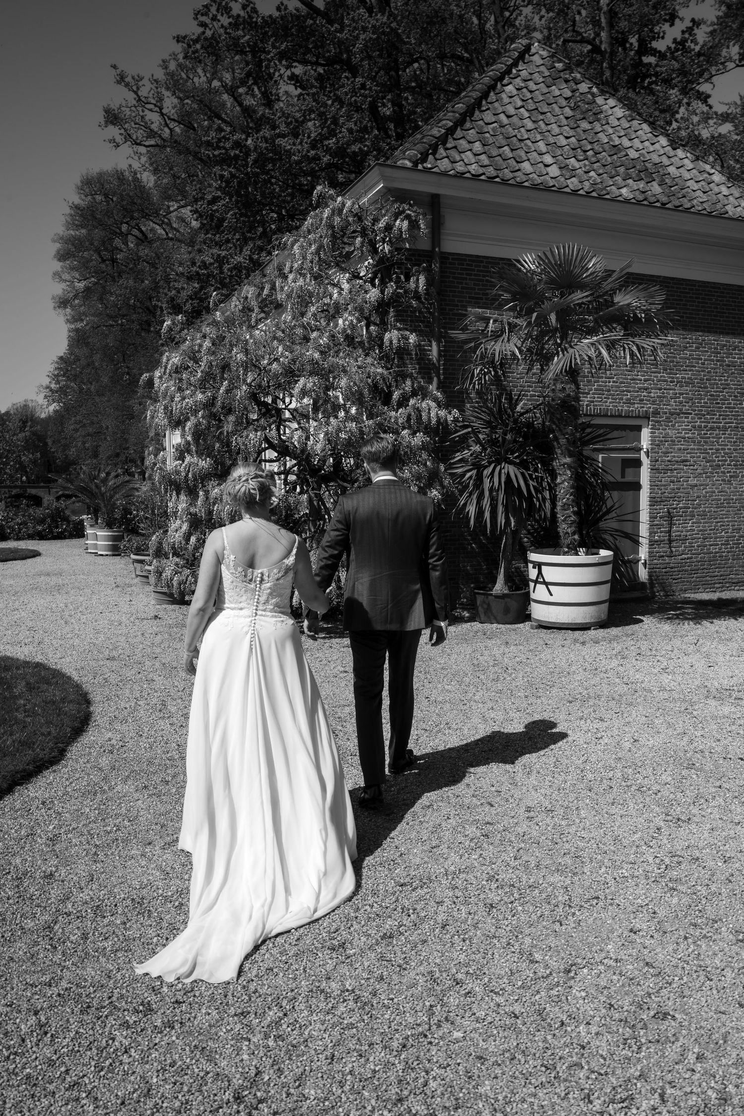 Black and white photo of bride and groom holding hands walking toward elegant estate surrounded by lush gardens.