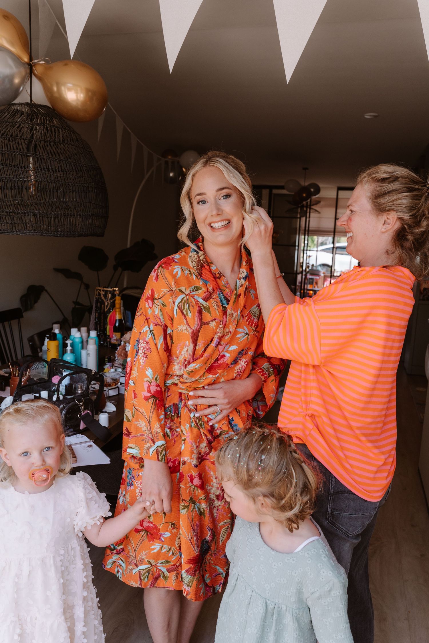Woman in orange floral robe gets earrings put on while two young children stand nearby at a festive indoor gathering.
