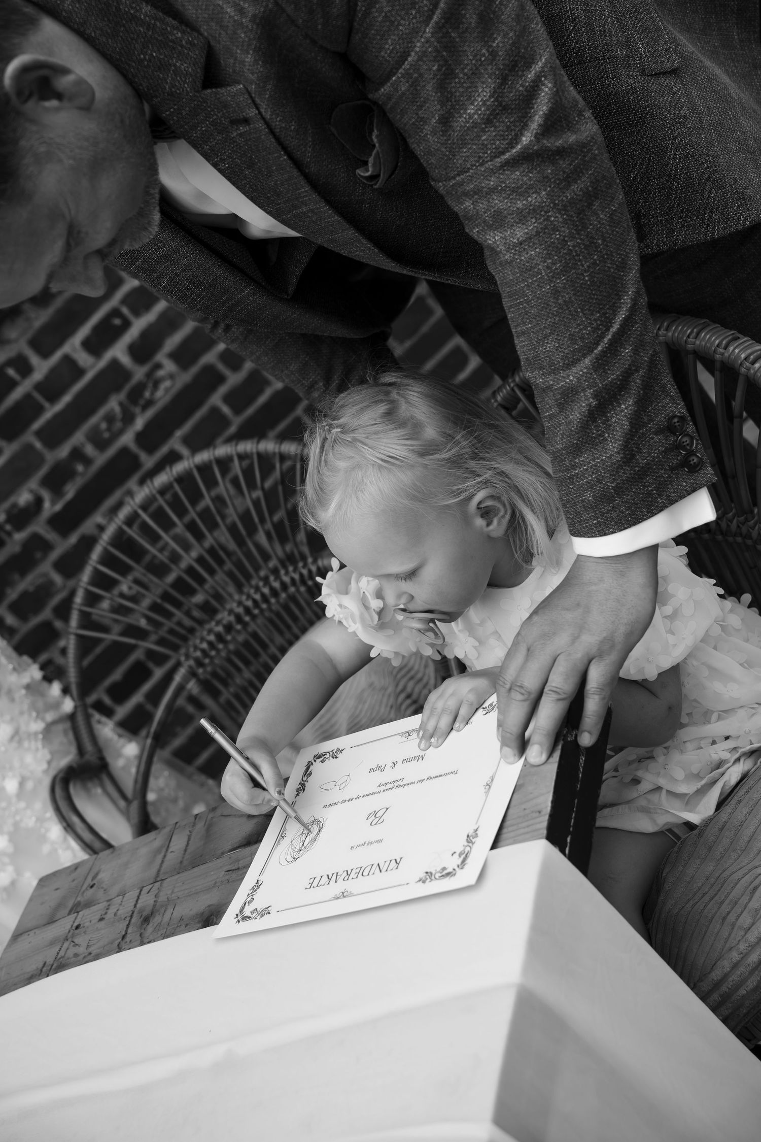 Black and white photo of a toddler signing a Kinderkarte certificate at a formal event, guided by an adult.